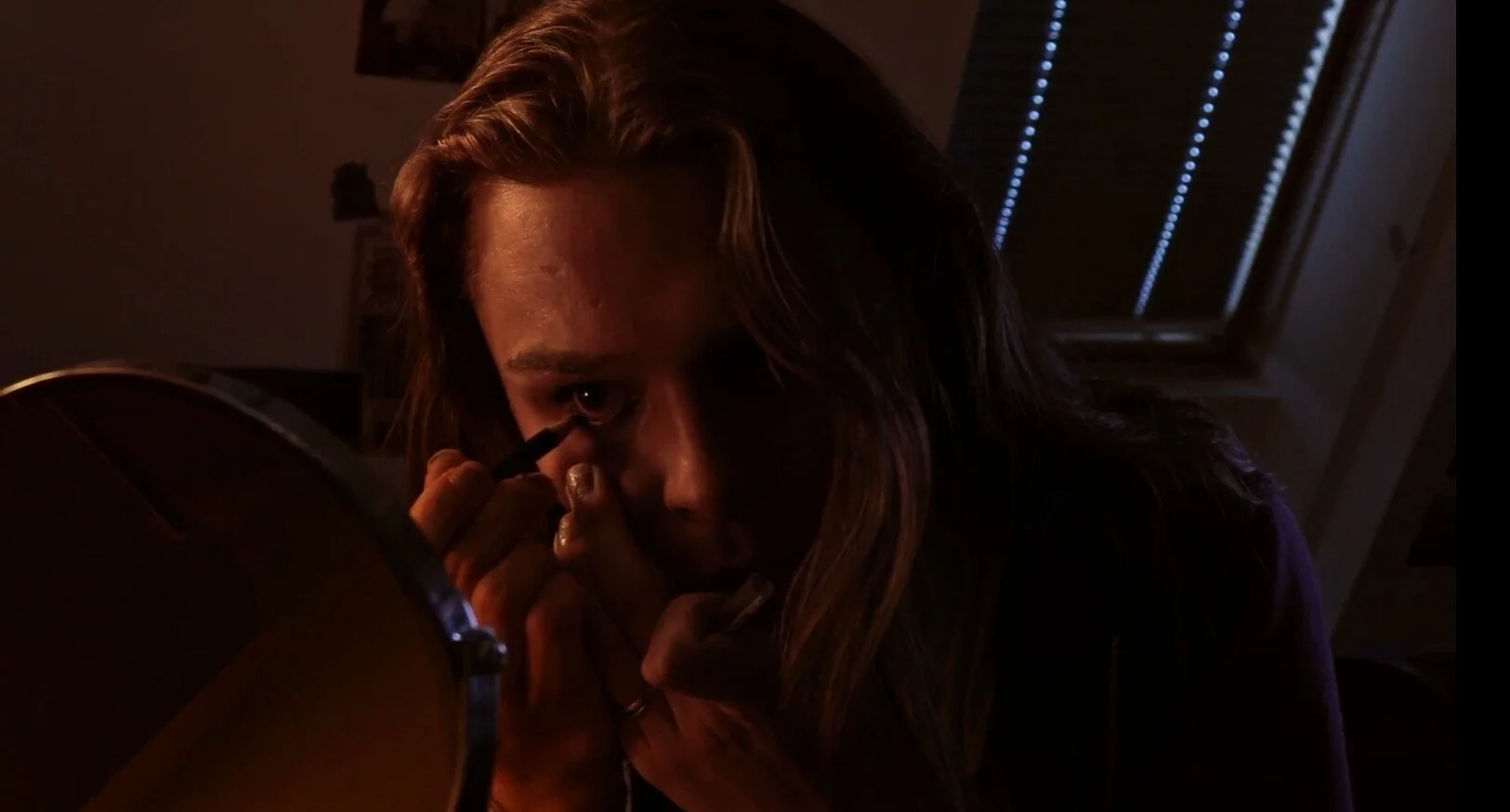 A woman with long hair applying makeup or eyeliner while looking into a mirror in a dimly lit room.