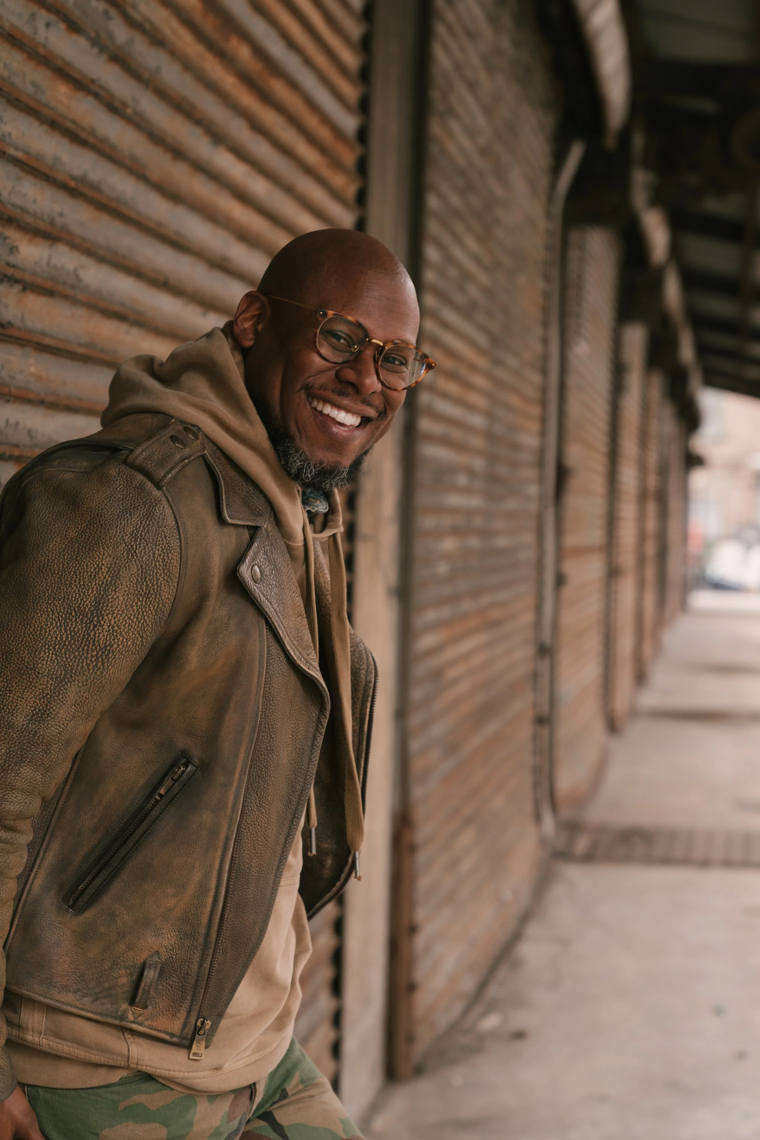 Smiling person wearing glasses and a leather jacket standing beside a rust-colored metal wall.