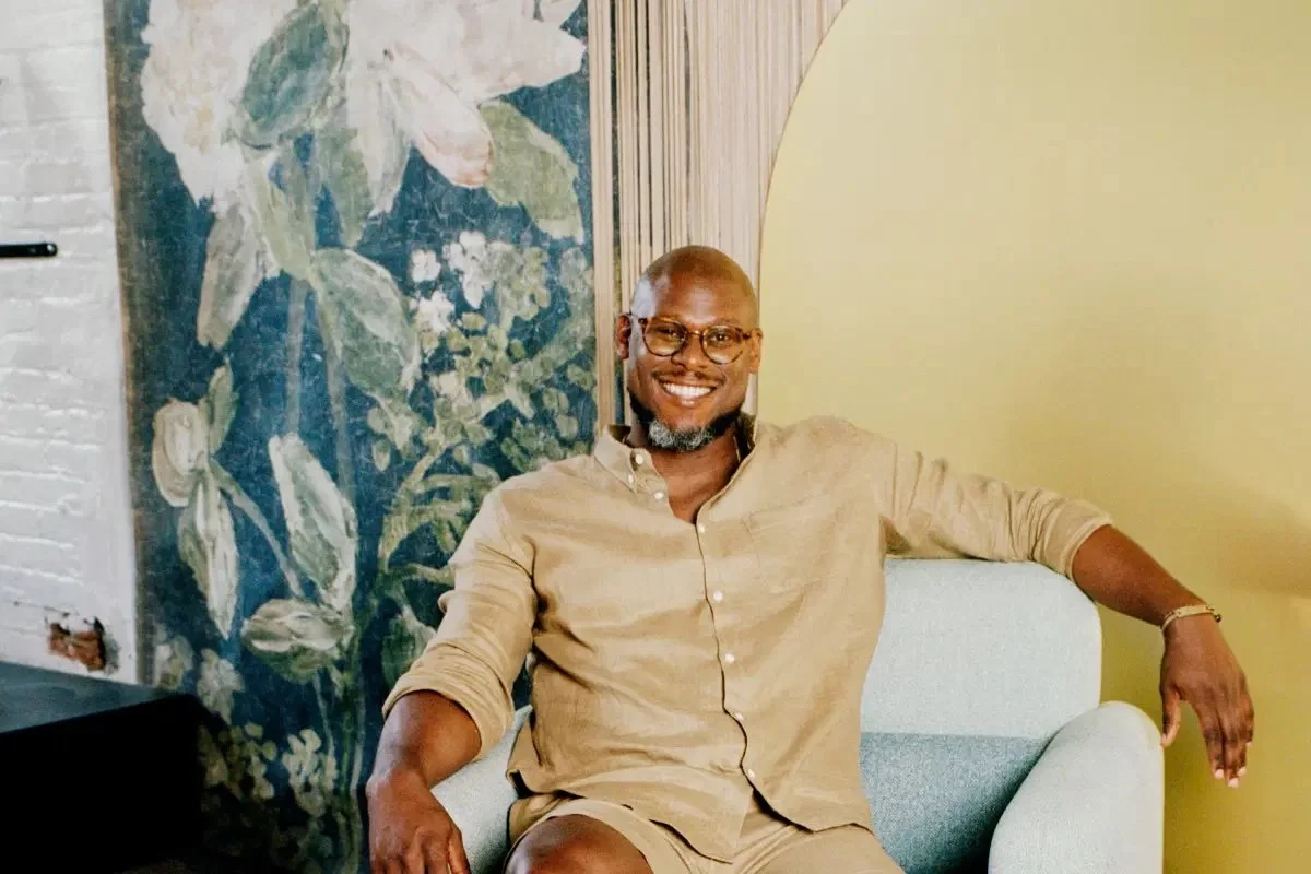 Person smiling while sitting on a light blue chair, wearing a beige shirt and shorts, with a floral mural and a yellow wall in the background.
