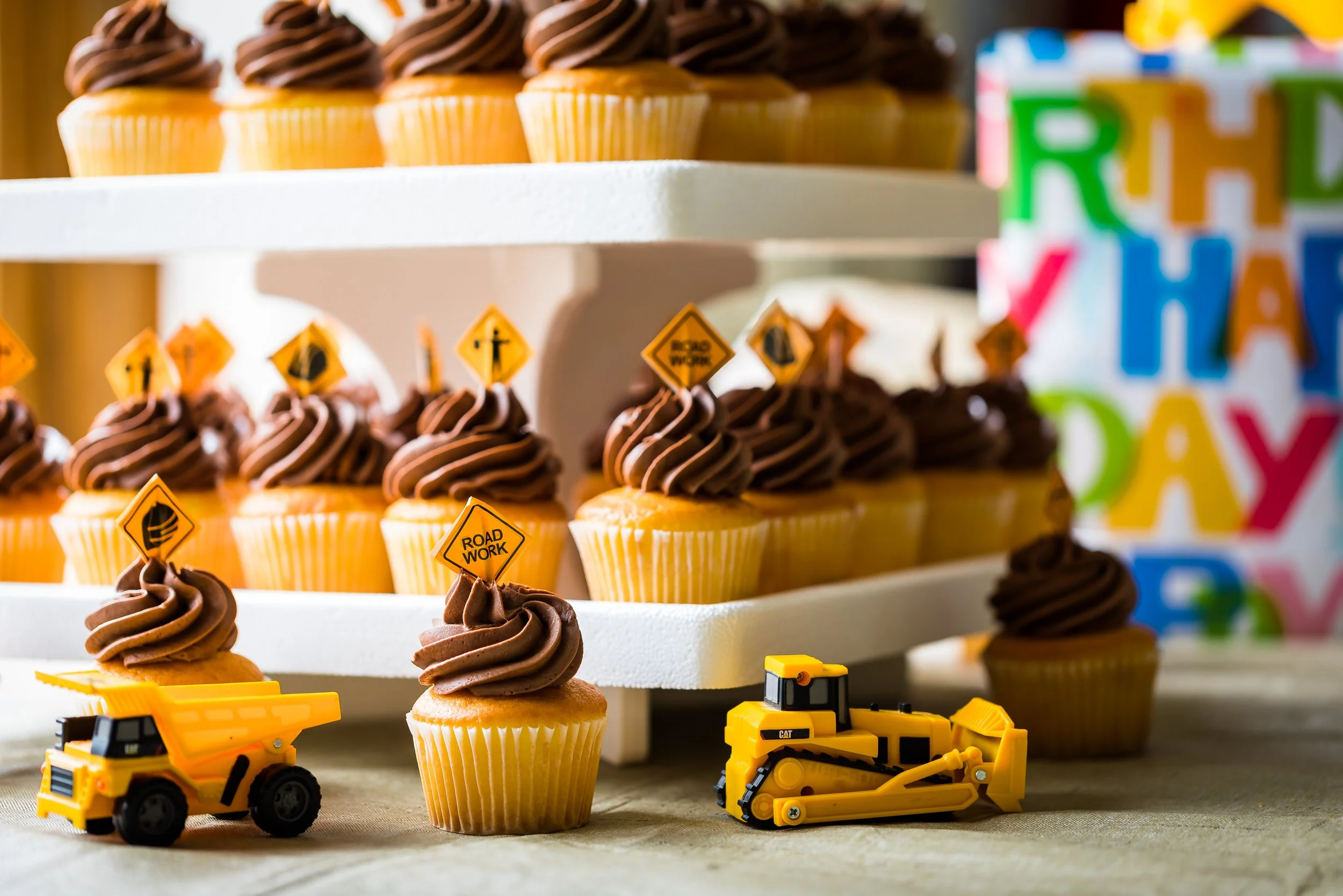 Cupcakes with chocolate frosting and toy construction vehicles, decorated with road work signs, on a stand near a colorful "Happy Birthday" background.