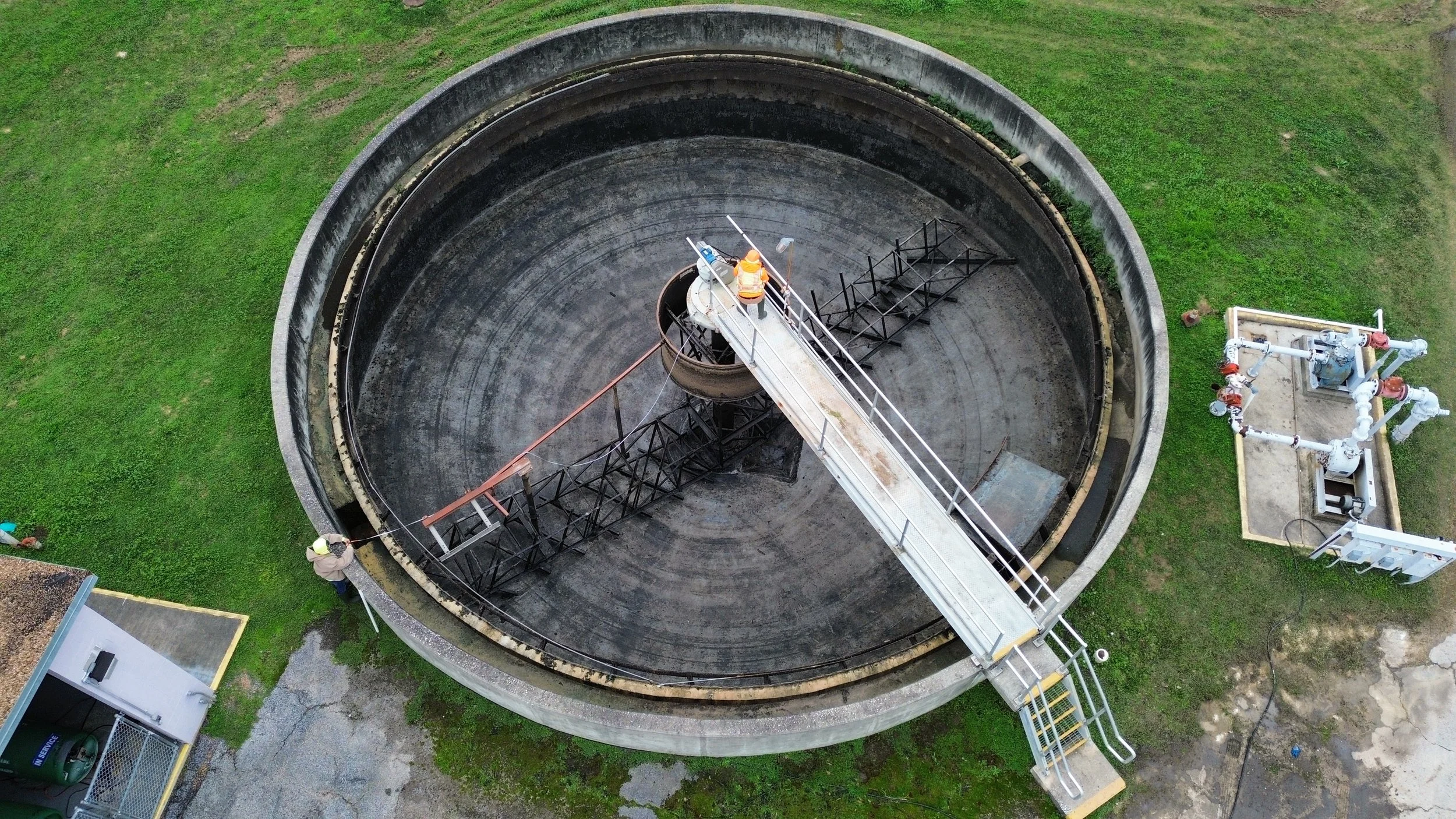 An aerial view of a circular water treatment tank with a worker on a lift inspecting inside, surrounded by green grass and some equipment.