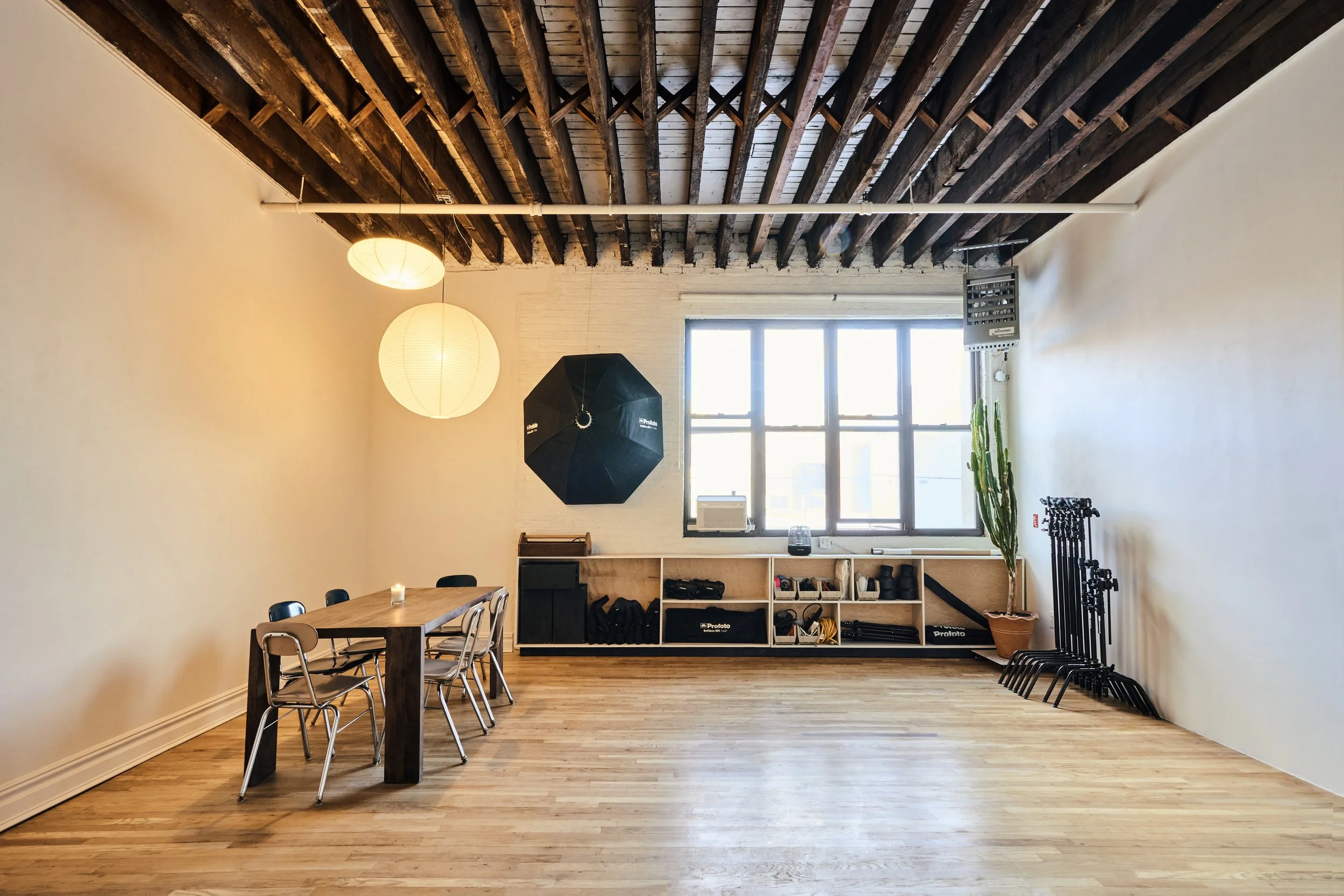 Spacious room with wooden floor, exposed beams, a long table with chairs, and photography equipment including a large umbrella light. Shelving units hold various gear, and there's a large window with natural light.