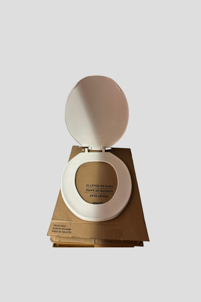 White toilet seat with lid open, placed on a cardboard box, with a blue storage container beneath, against a gray striped wall.