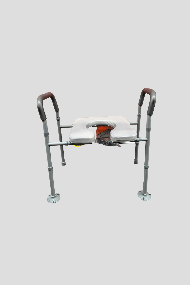 A hospital shower chair with a white cushioned seat and backrest, gray frame, and black rubber armrests, placed on a multicolored speckled floor in front of a bright red corrugated metal wall.