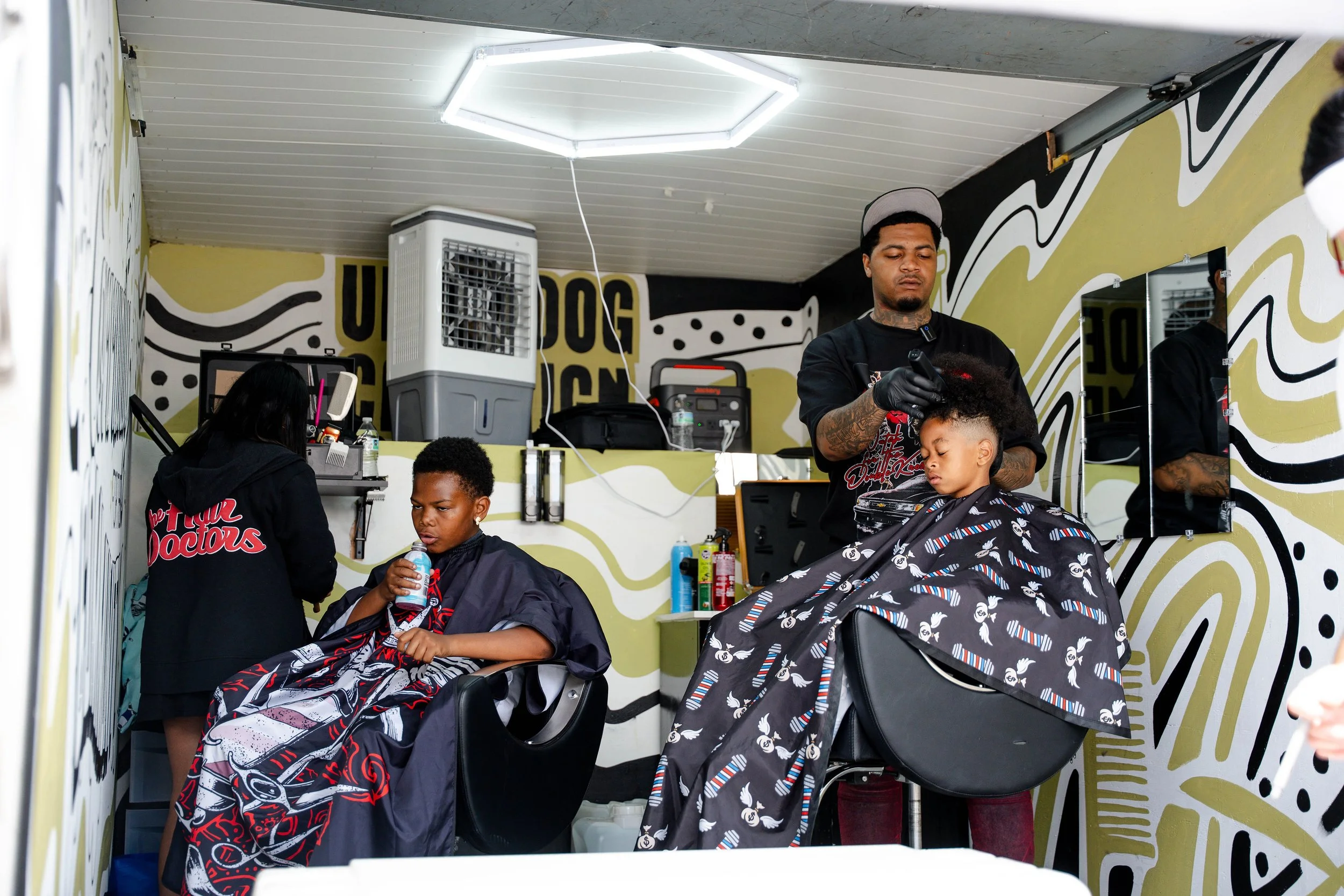 Barbershop scene with a barber cutting a young man's curly hair, another young man sitting with a cape, and a woman in the background inside a decoratively painted room.