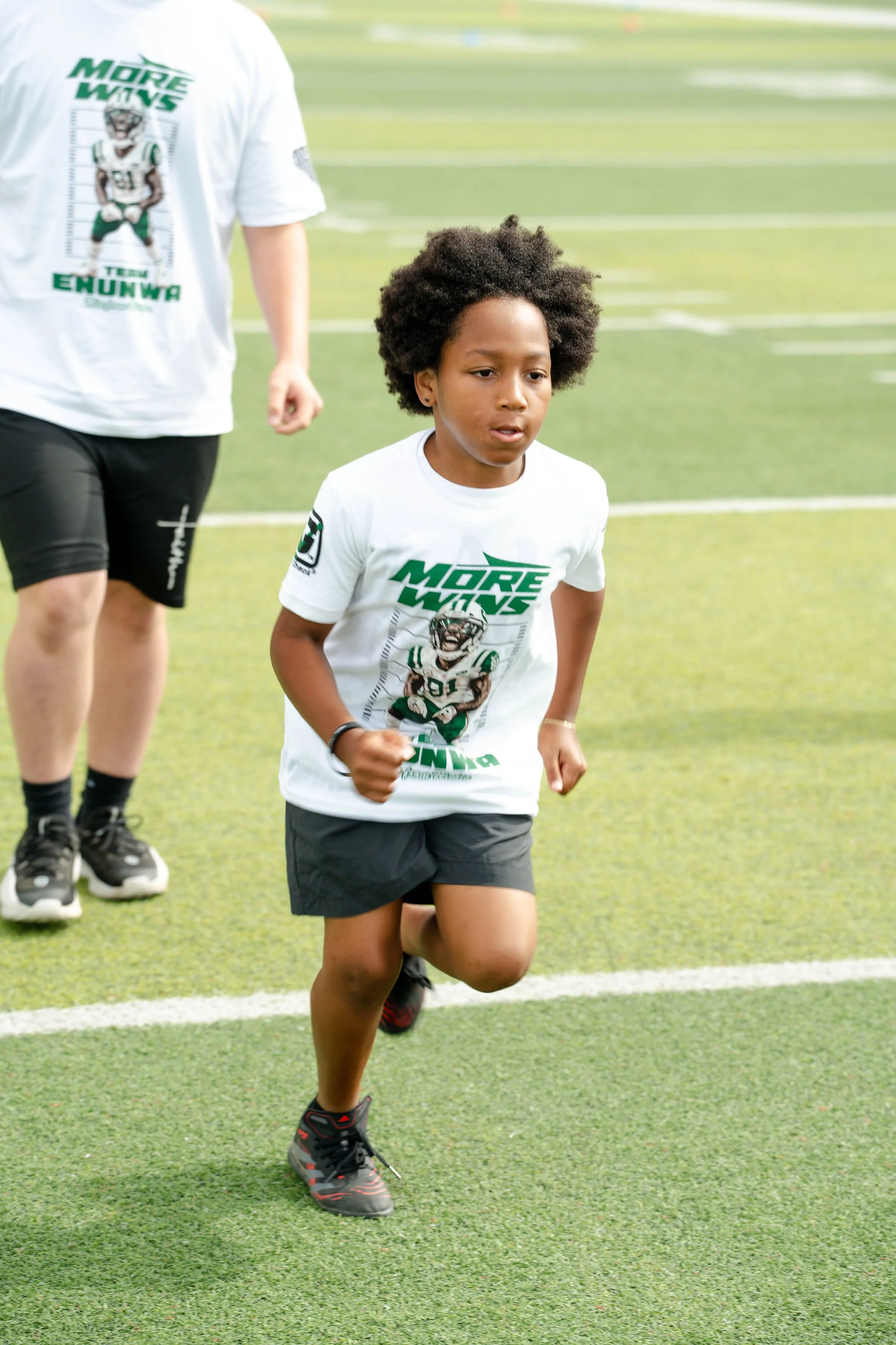 Young girl running on a green sports field wearing a white T-shirt with sports-themed text and graphics, black shorts, and black sneakers with red accents.