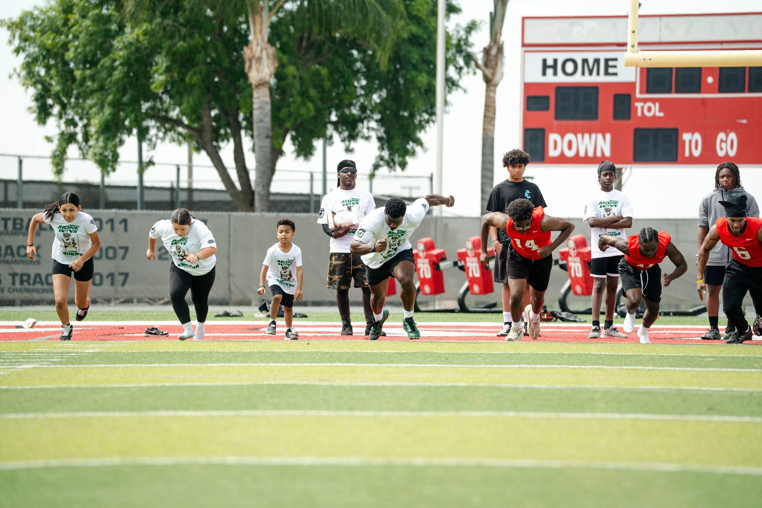 Kids and adults practicing running drills on a sports field, with a scoreboard and trees in the background.