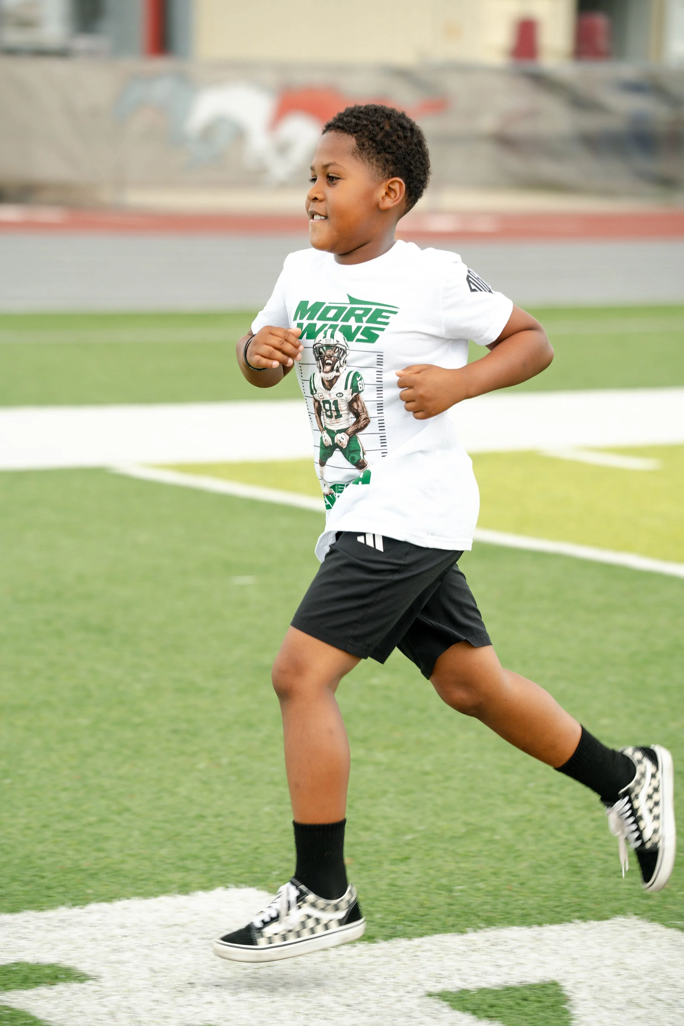 A young boy running on a sports field, wearing a white T-shirt with a graphic design and the words 'More Wins,' black shorts, black socks, and checkered sneakers.