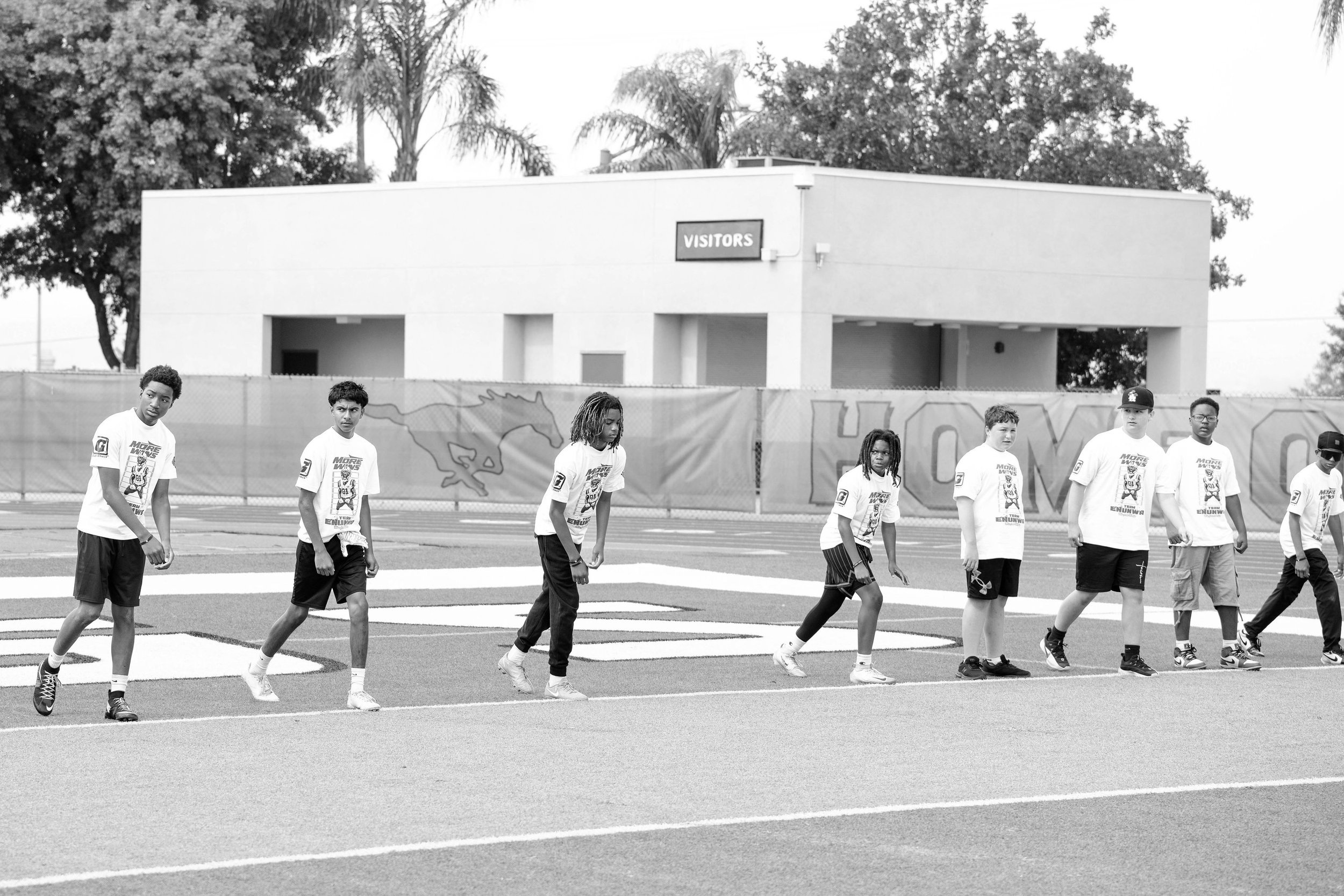 Group of young boys in white T-shirts and shorts walking on a sports field, with a building labeled 'Visitors' and trees in the background.