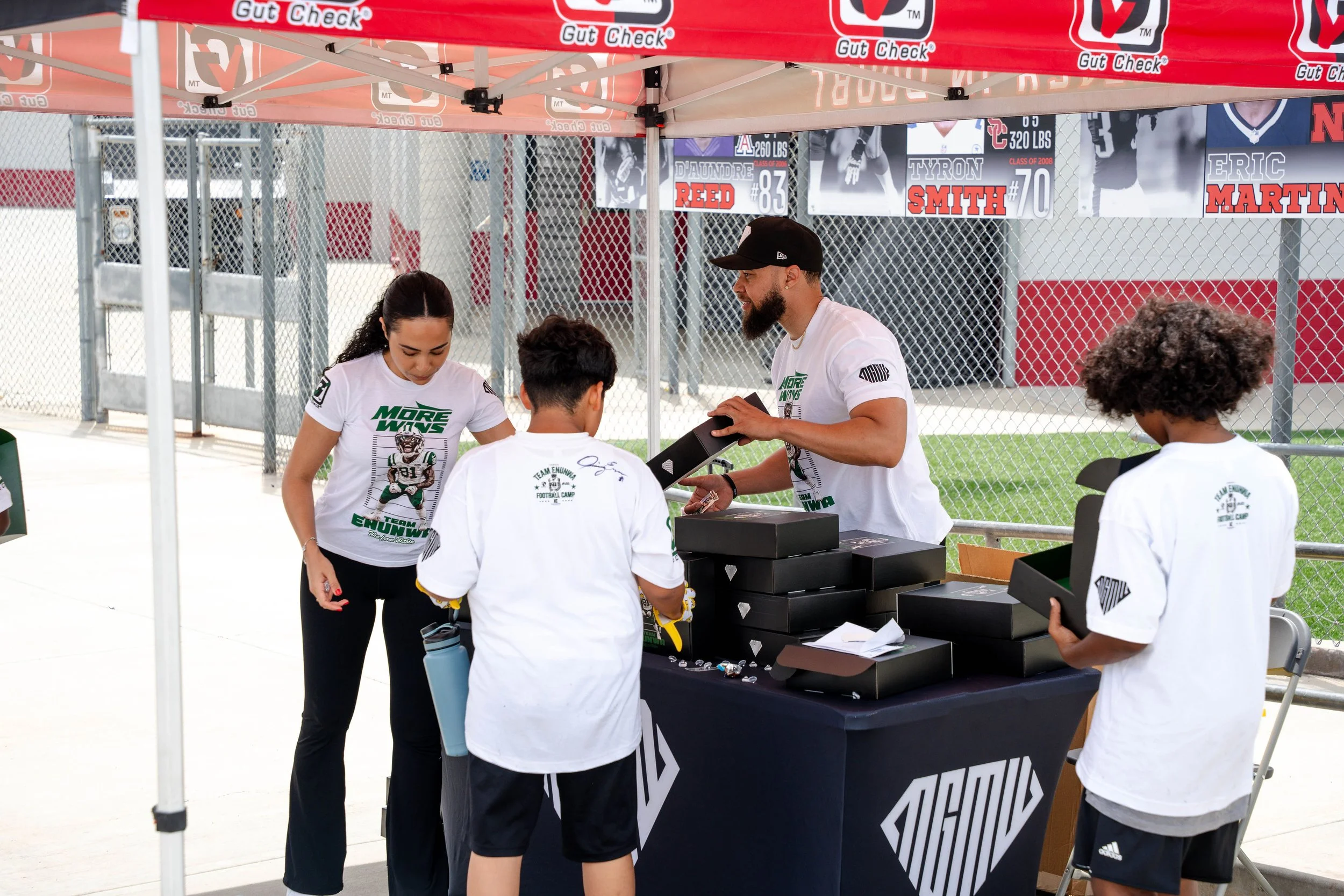 People gathered at a registration or merchandise table under a red tent with 'Gut Check' branding at an outdoor sports event, with chain-link fencing and sports banners in the background.