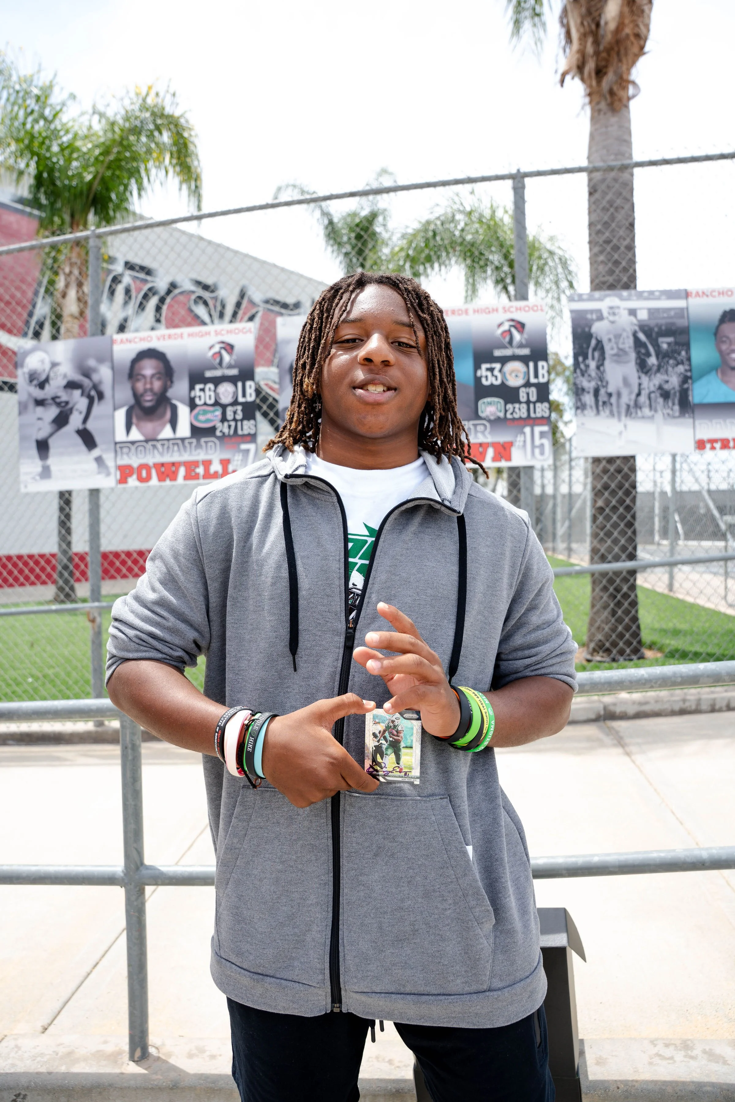 Young man with dreadlocks wearing a gray hoodie and multiple wristbands standing outside in front of a chain-link fence with photos of football players and banners for Rancho Verde High School in the background, palm trees visible behind the fence.