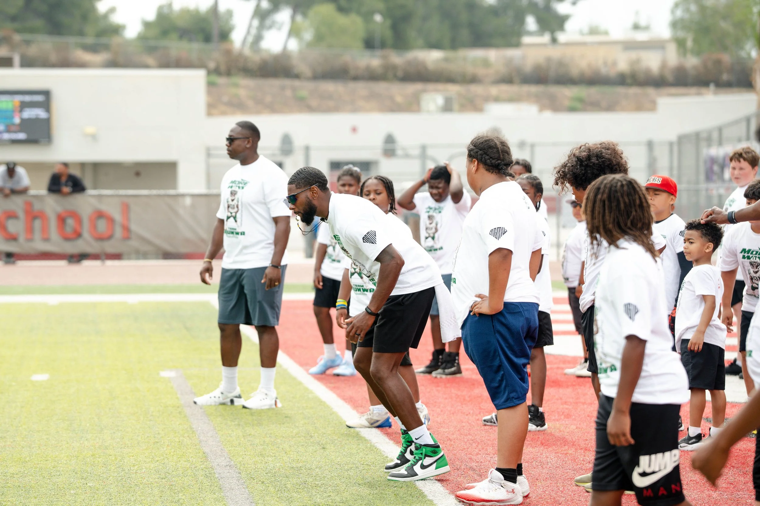 A group of children and adults on a sports field, participating in an outdoor athletic activity, with some wearing athletic gear and trainers, and a stadium with a digital scoreboard in the background.