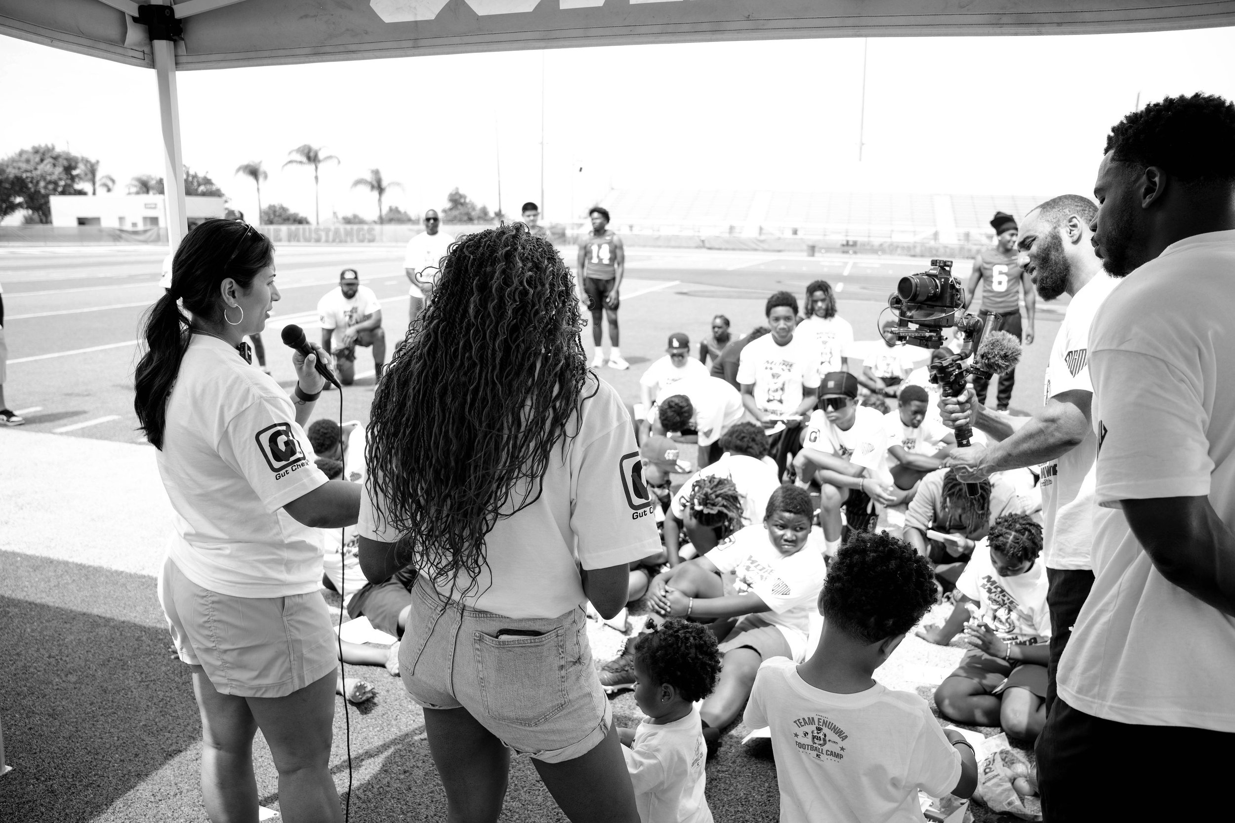 A group of children and adults gathered outdoors on a football field for an event. Some are sitting on the ground, and others are standing and talking. The background features bleachers, palm trees, and a clear sky.