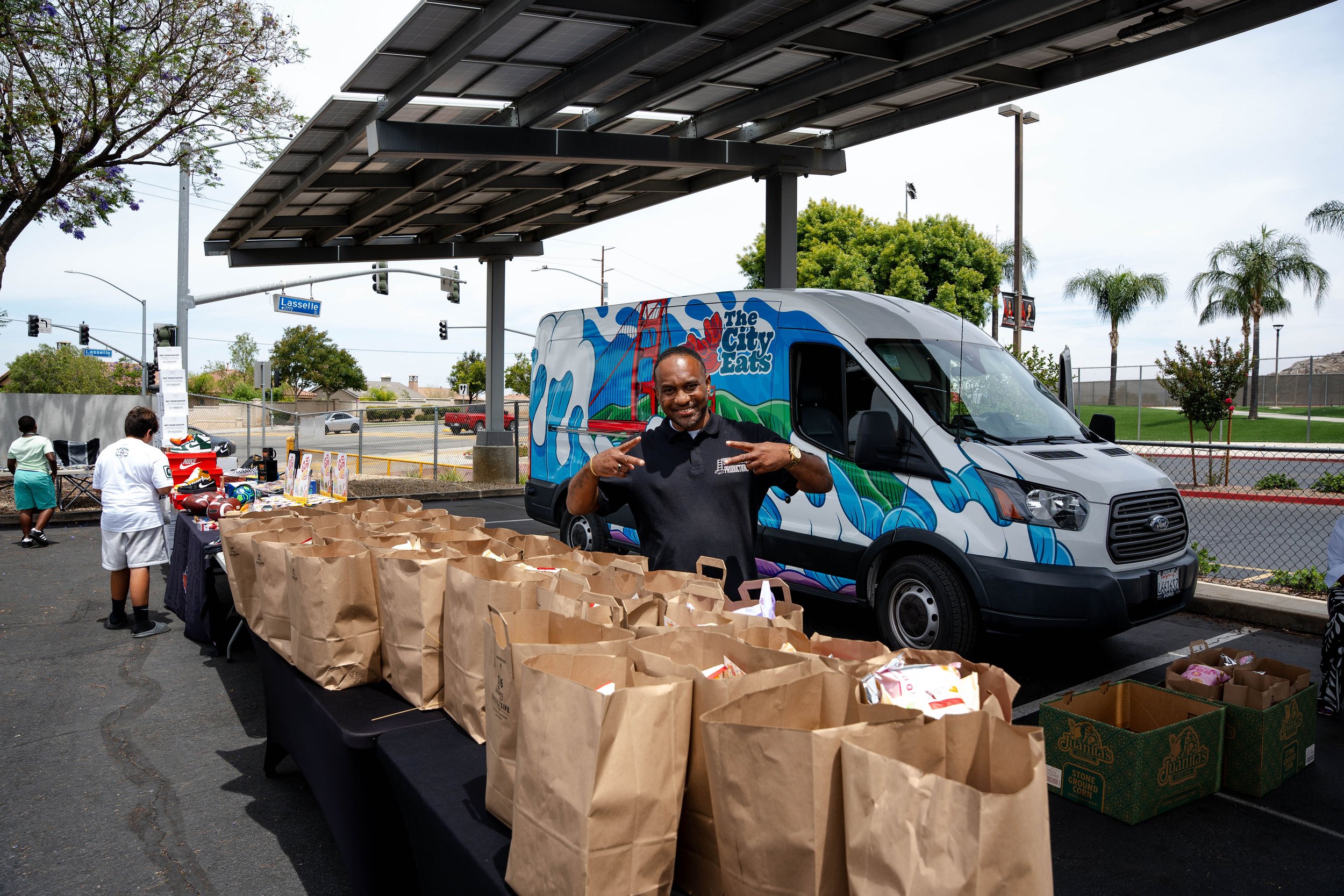 Man standing in front of a van with the logo "The City Eats" under a solar panel structure, with tables of brown paper bags containing food for distribution and people in the background at an outdoor event.