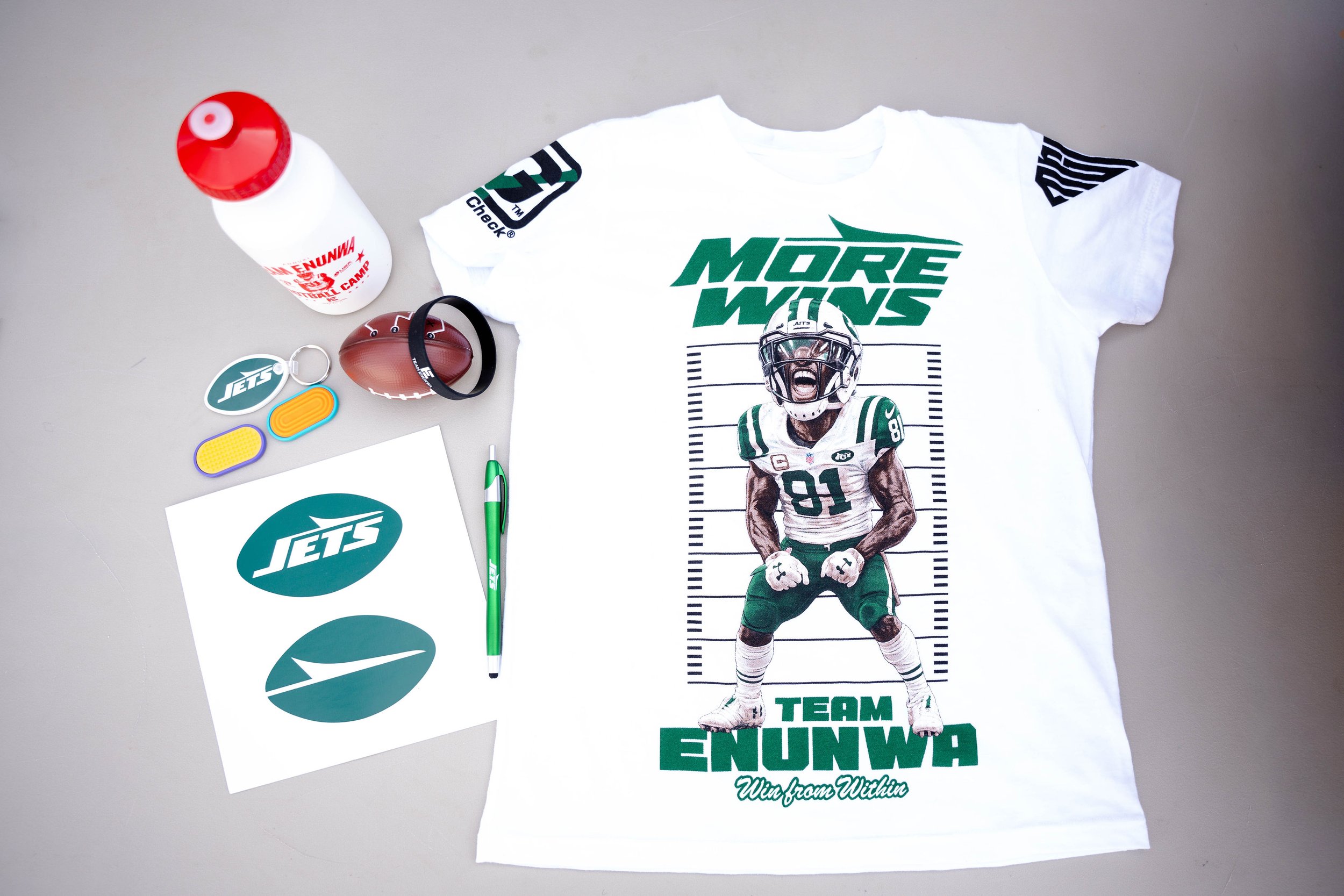 A white t-shirt with bold green text that says 'MORE WINS' and an image of a football player in a green uniform with a helmet, underneath the text it says 'TEAM ENUNWA' and 'Win from Within'. Next to the t-shirt are various items including a white wa