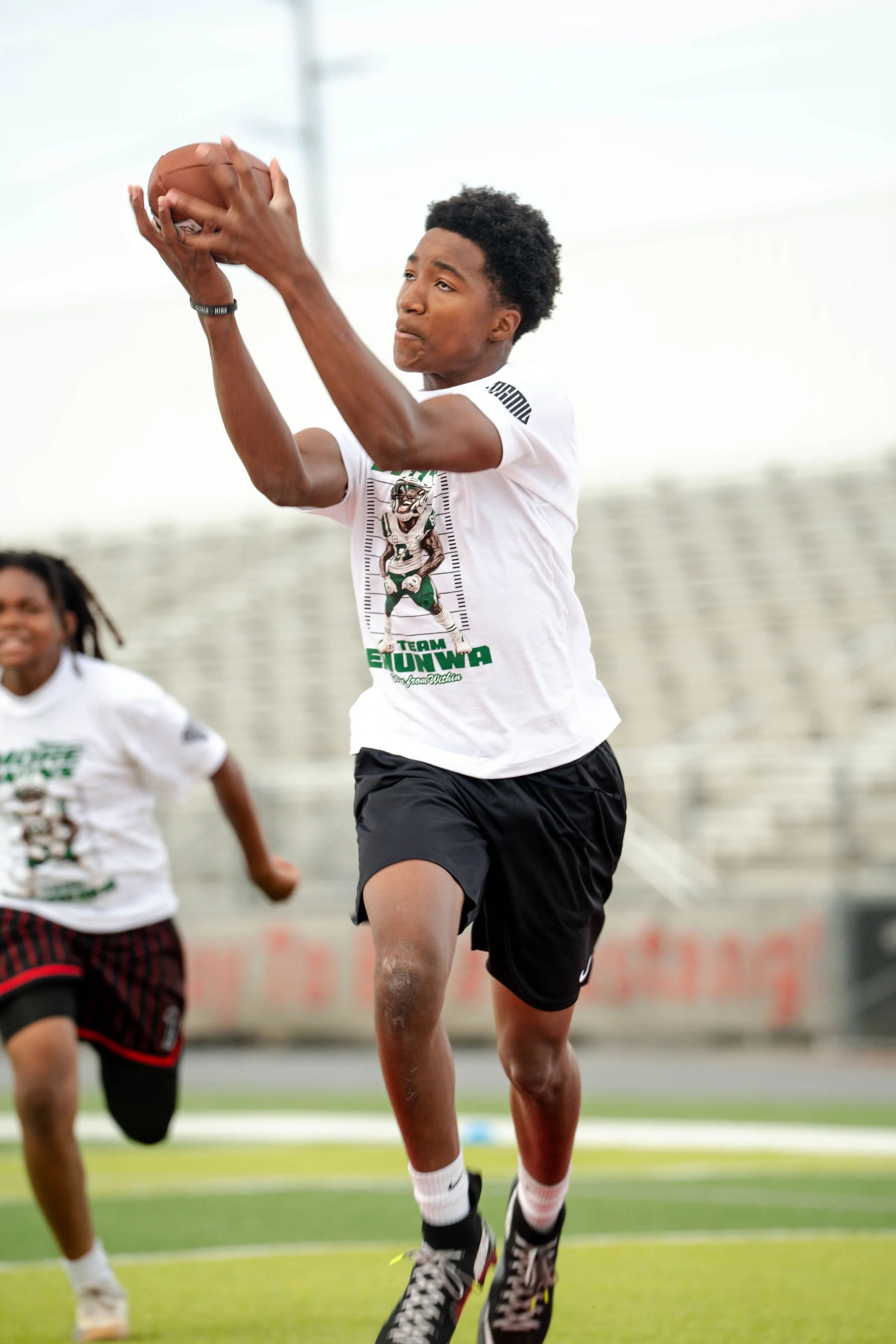 A young athlete catching a football during practice on a sports field, with another player running nearby.