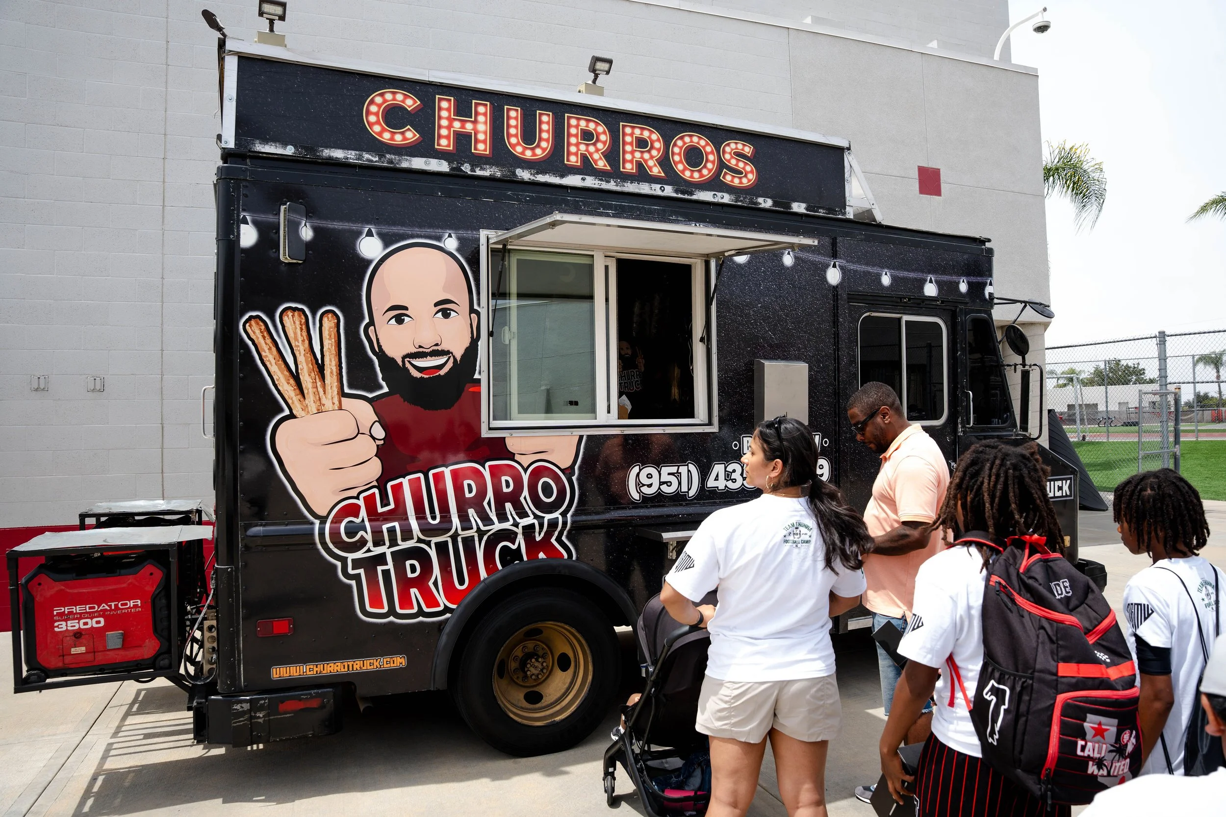 A black food truck named 'Churros Truck' with a large sign and a cartoon illustration of a man holding churros. Several people, including children in white shirts and backpacks, are standing in line around the truck outdoors near a fence and a gray b