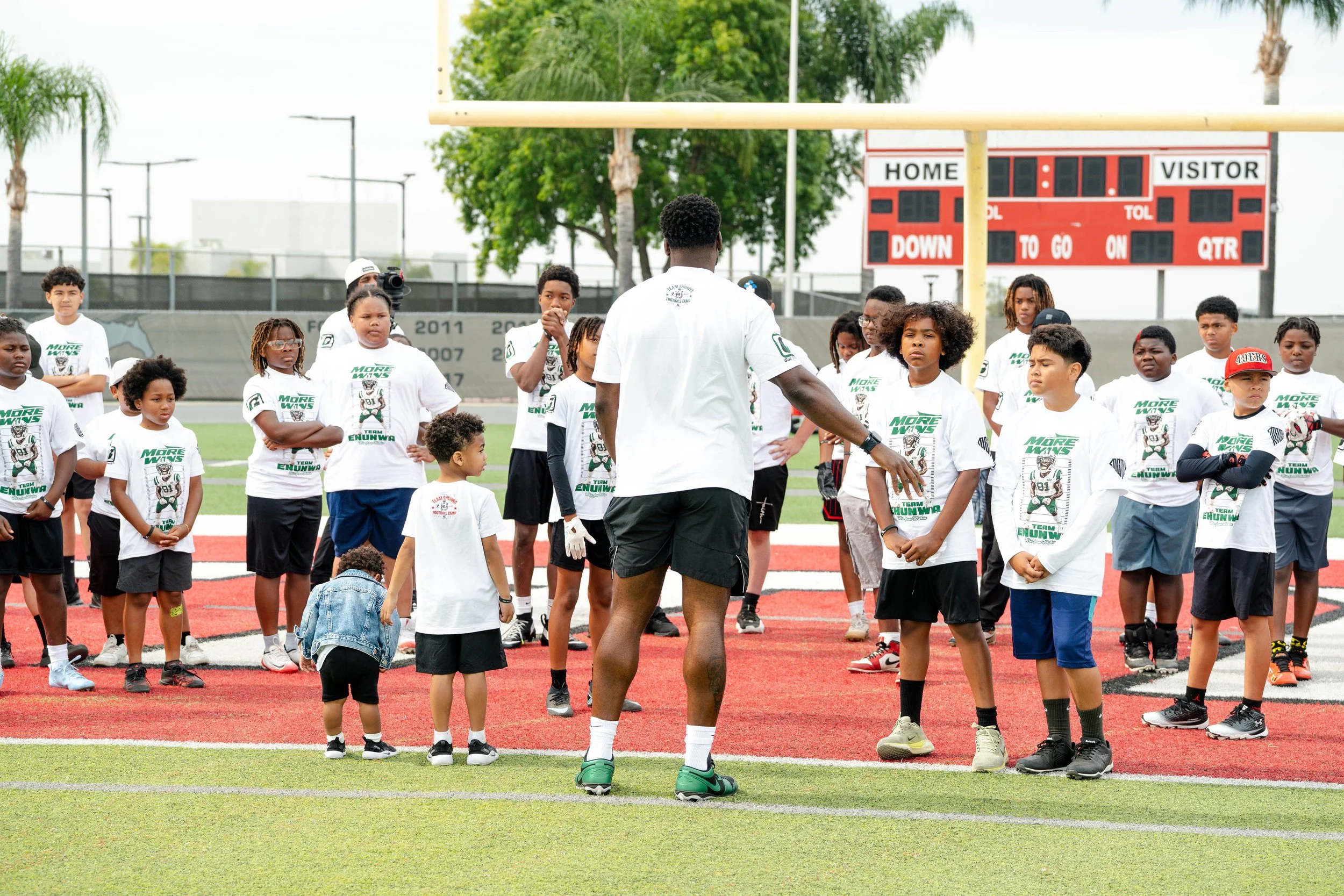 A group of children and a coach gathered on a football field during a practice session. The children are wearing white T-shirts with the words 'More Wins' and a cartoon mascot printed on them, standing in front of a goalpost. The coach, with his back