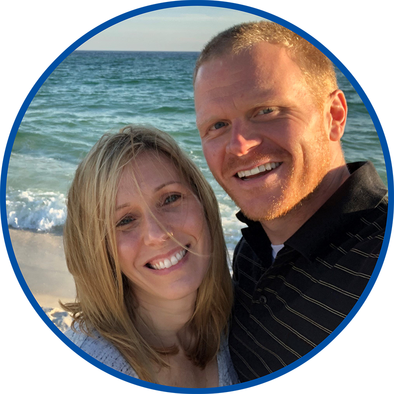 A couple smiling and taking a selfie on a beach with the ocean in the background.