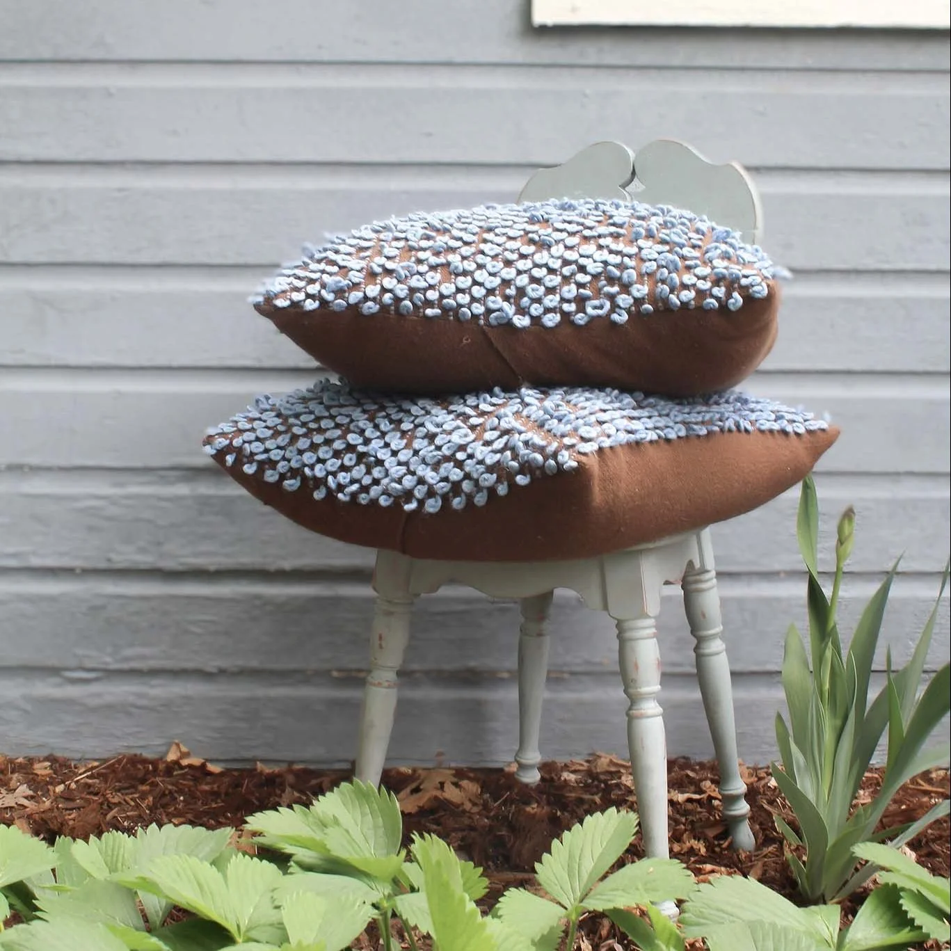 Two brown pillows with white and blue details stacked on a small white painted wooden chair outside, surrounded by plants, in front of a grey wooden wall.