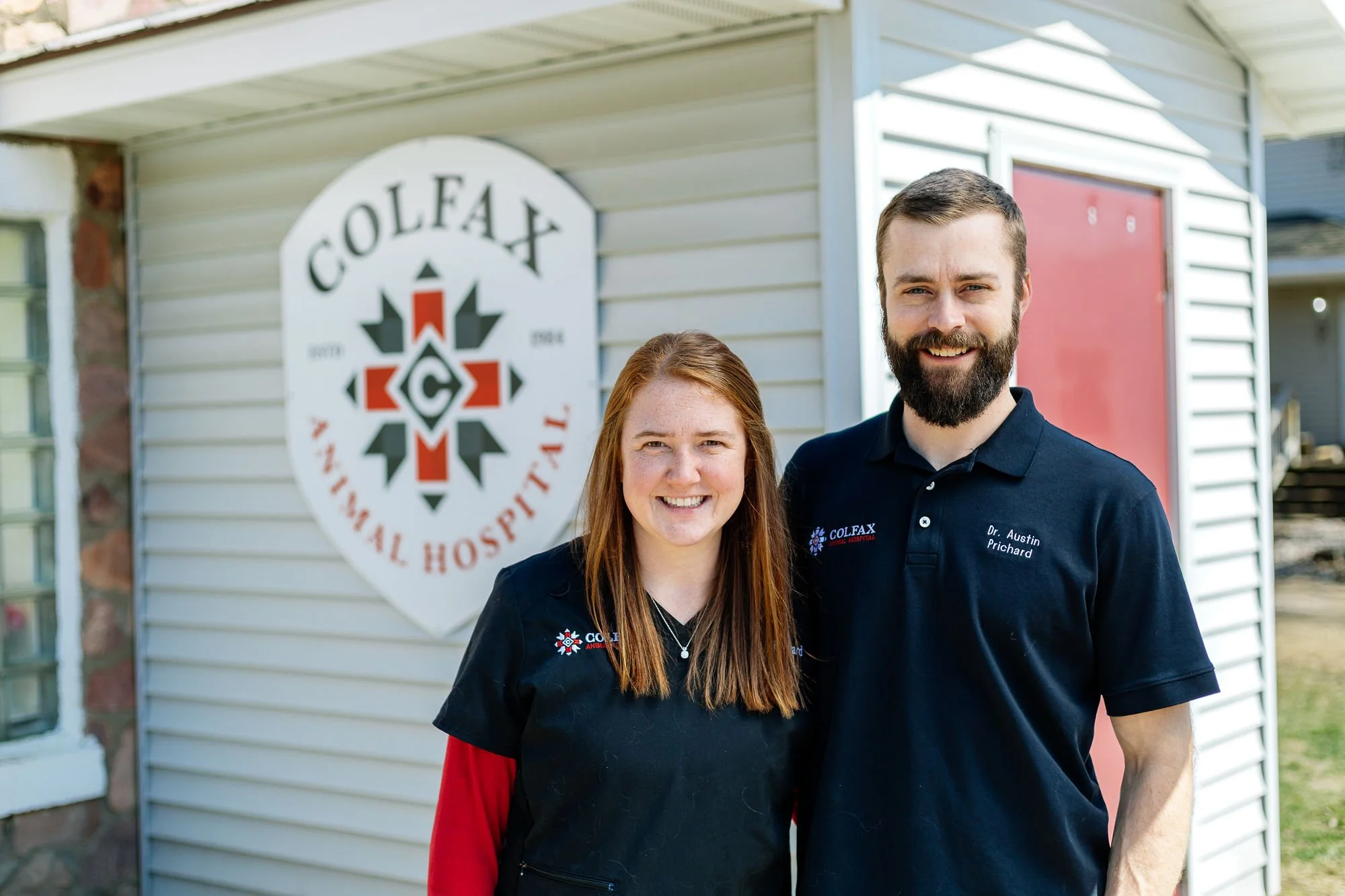 Two people standing outside in front of a building with a sign that says 'COLFAX ANIMAL HOSPITAL' and a logo. One is a woman with long red hair wearing a black jacket and red shirt. The other is a man with a beard and short hair wearing a black polo shirt with embroidery.