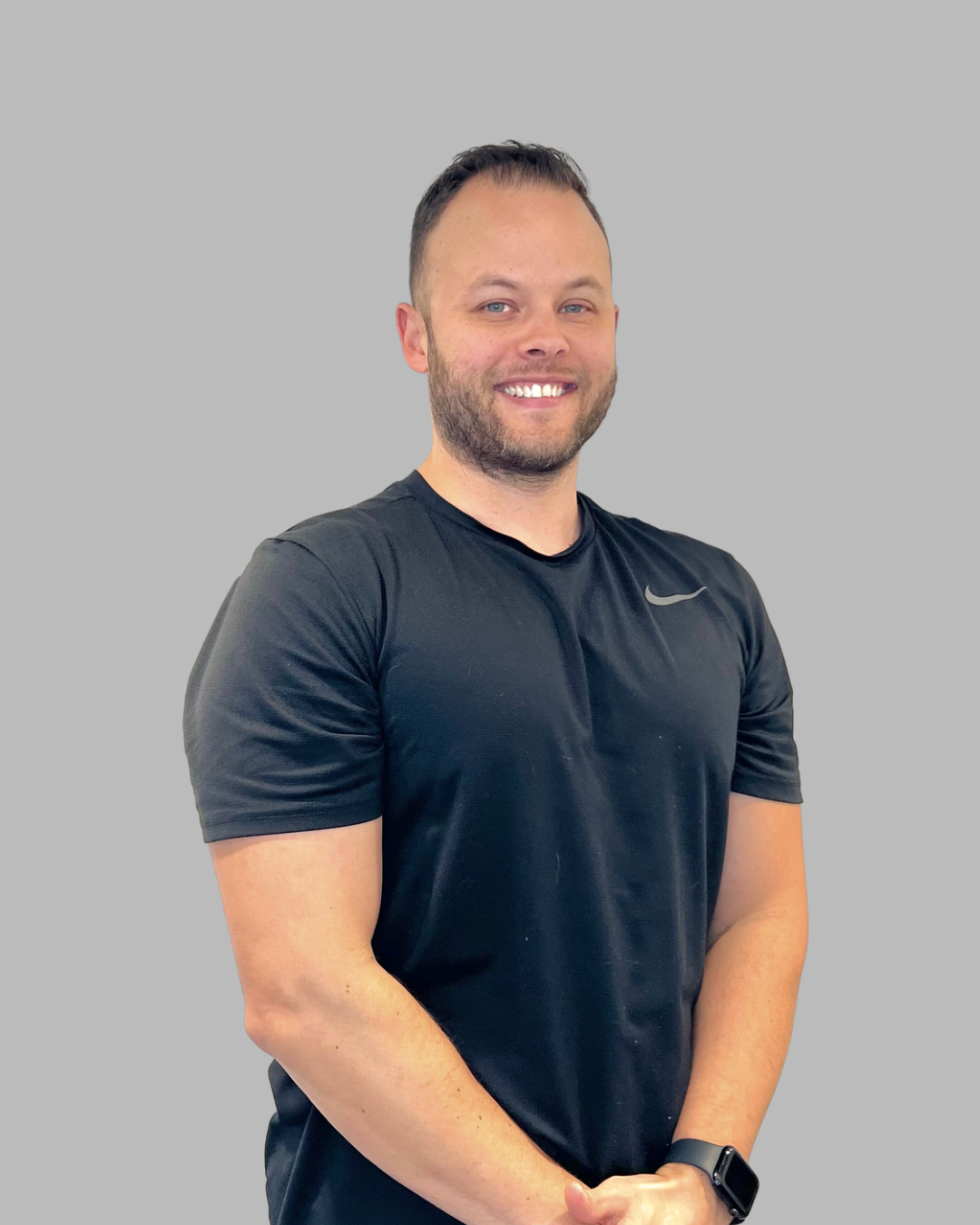 A smiling man with short dark hair and a beard, wearing a black Nike athletic shirt and a smartwatch, standing against a plain light-colored wall.