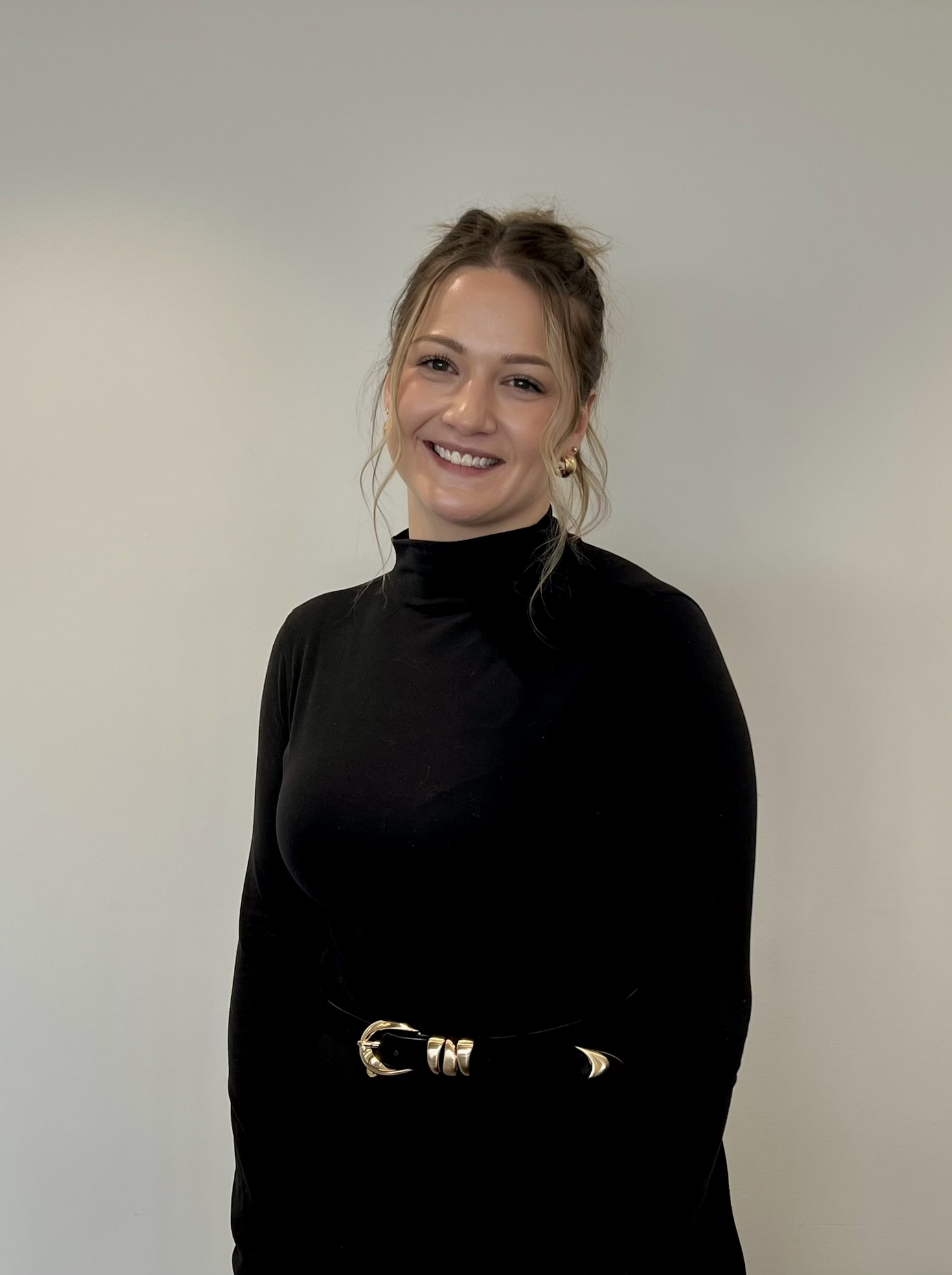 A young woman with long wavy blonde hair, smiling, wearing a gray sleeveless sweater over a black turtleneck, and a delicate necklace, standing in front of a plain off-white wall.
