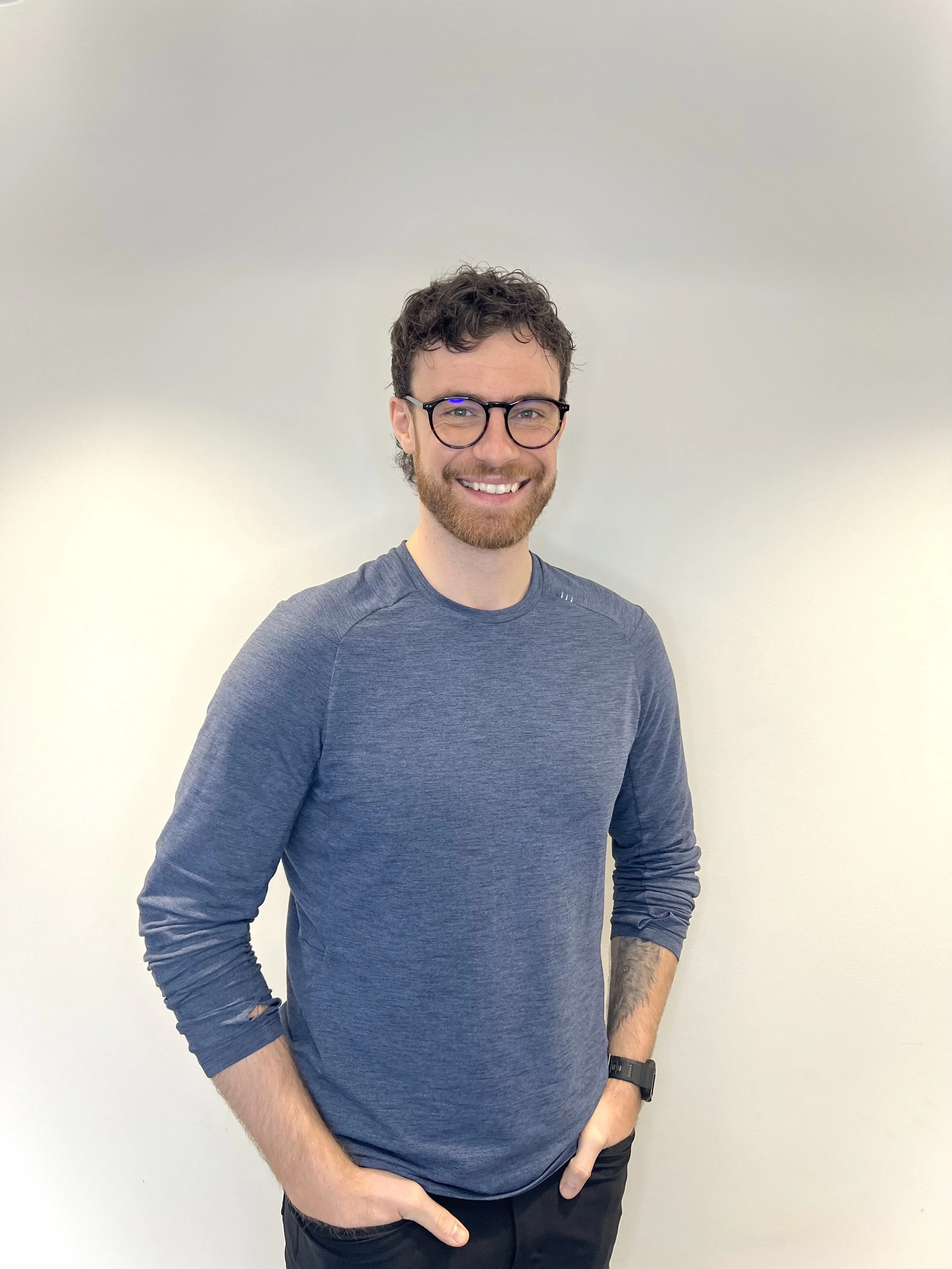 A smiling man with curly brown hair, glasses, and a beard, wearing a blue long-sleeve shirt and black pants, standing against a plain light-colored wall.
