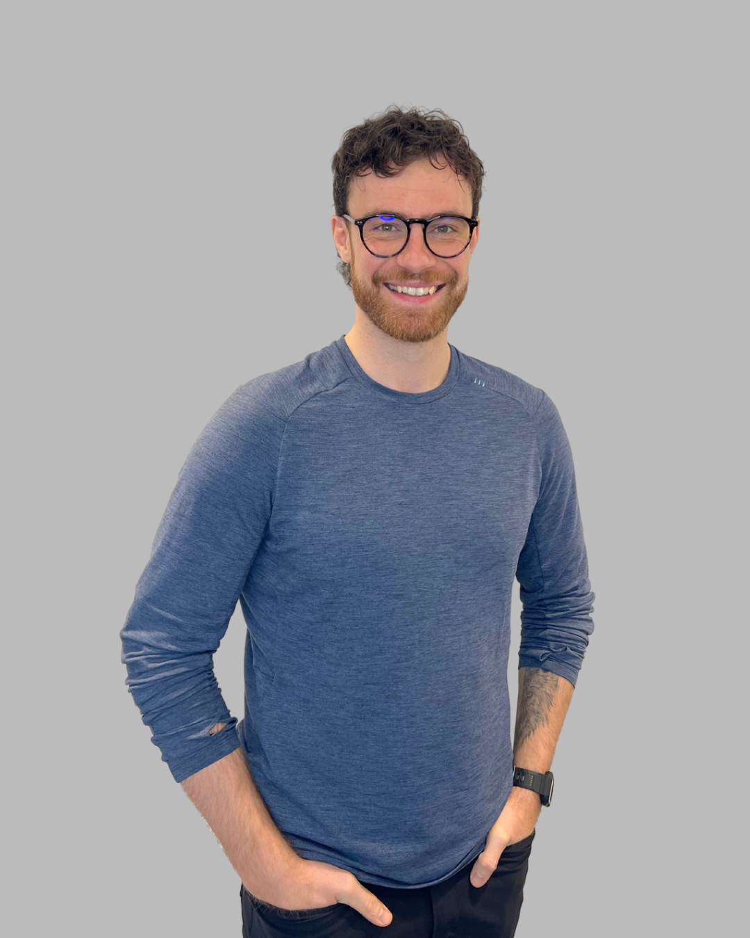 A smiling man with curly brown hair, glasses, and a beard, wearing a blue long-sleeve shirt and black pants, standing against a plain light-colored wall.