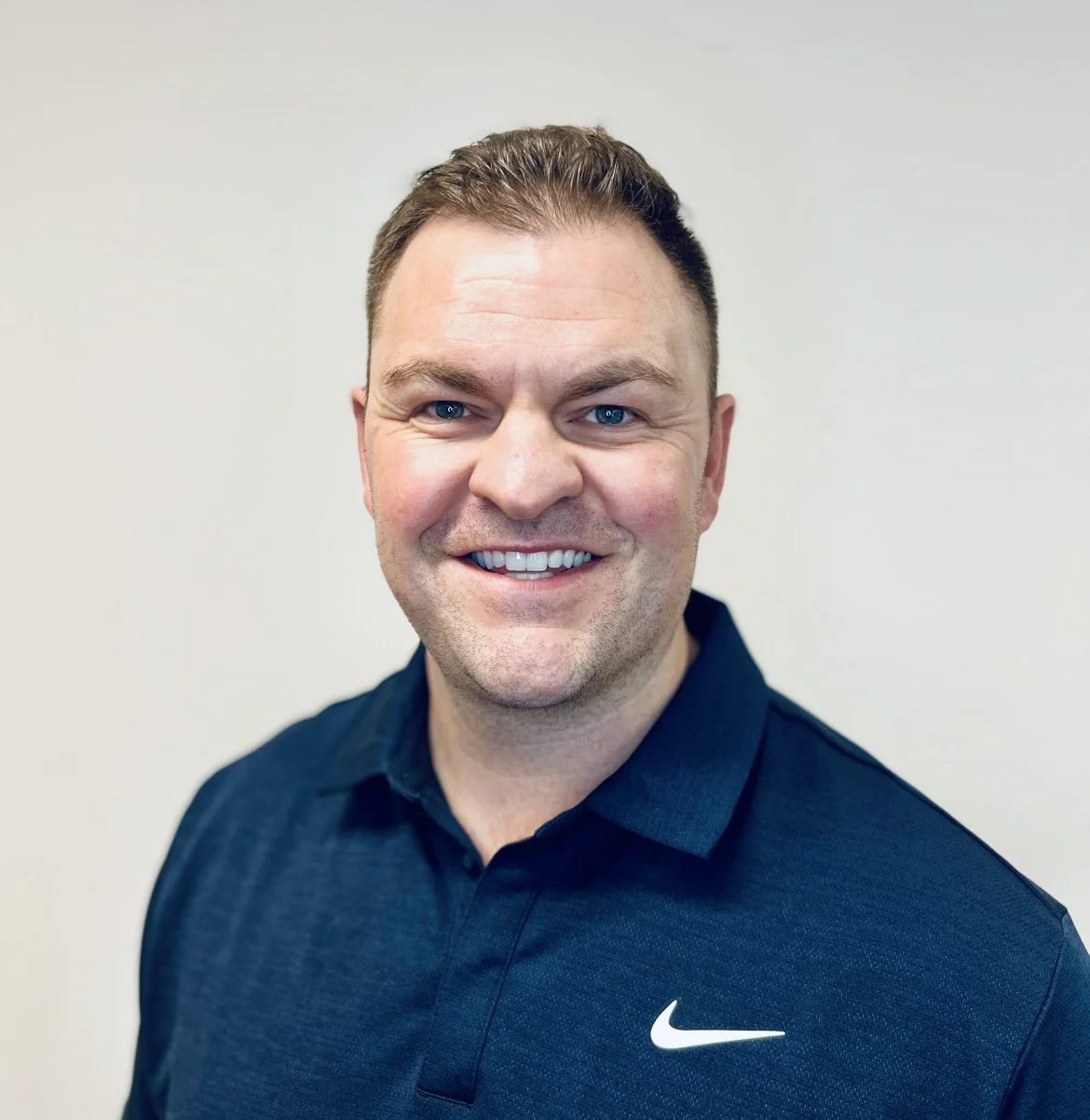 Headshot of a smiling man with short brown hair, blue eyes, wearing a black Nike polo shirt, standing against a plain light-colored background.
