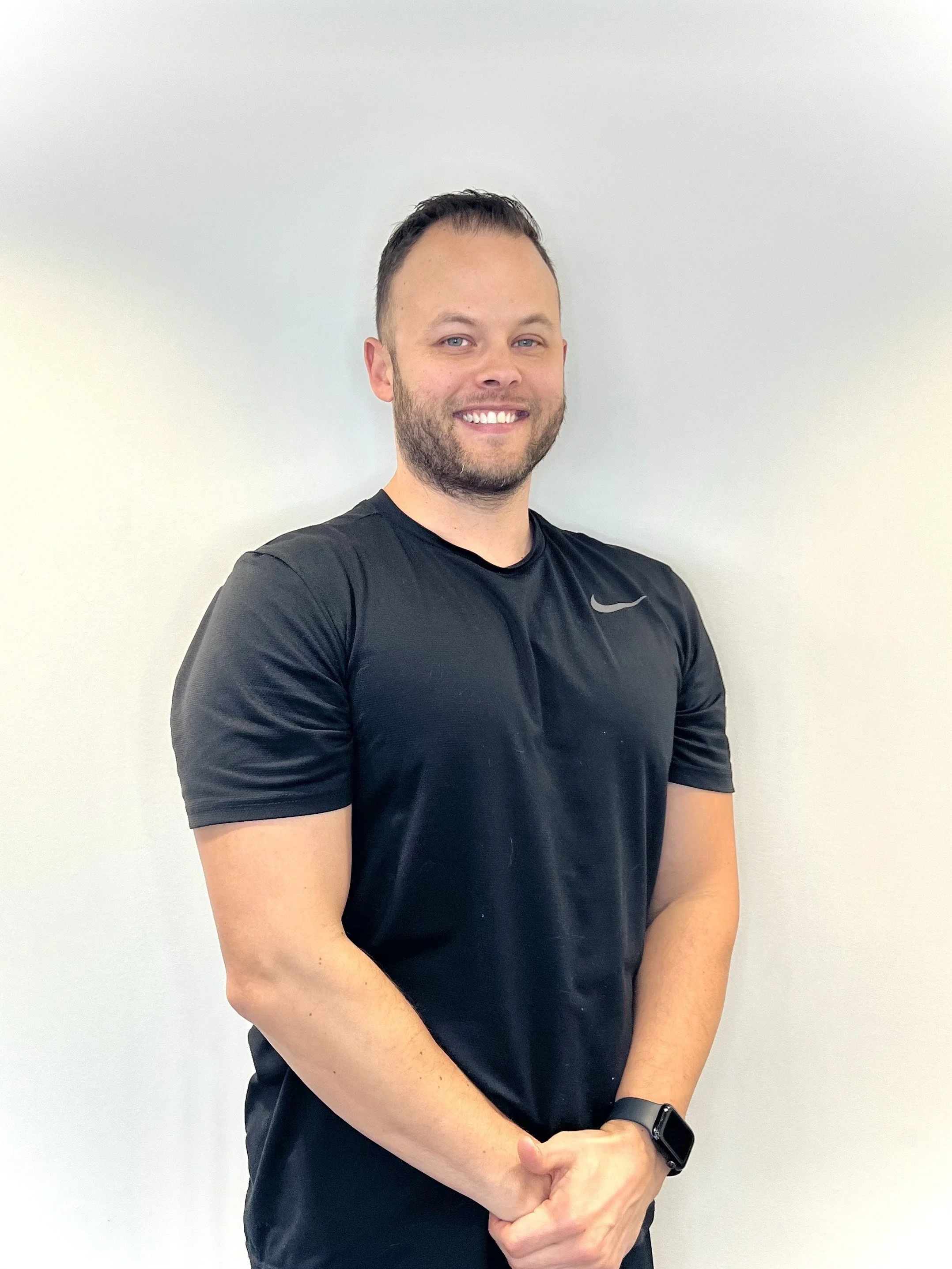 A smiling man with short dark hair and a beard, wearing a black Nike athletic shirt and a smartwatch, standing against a plain light-colored wall.