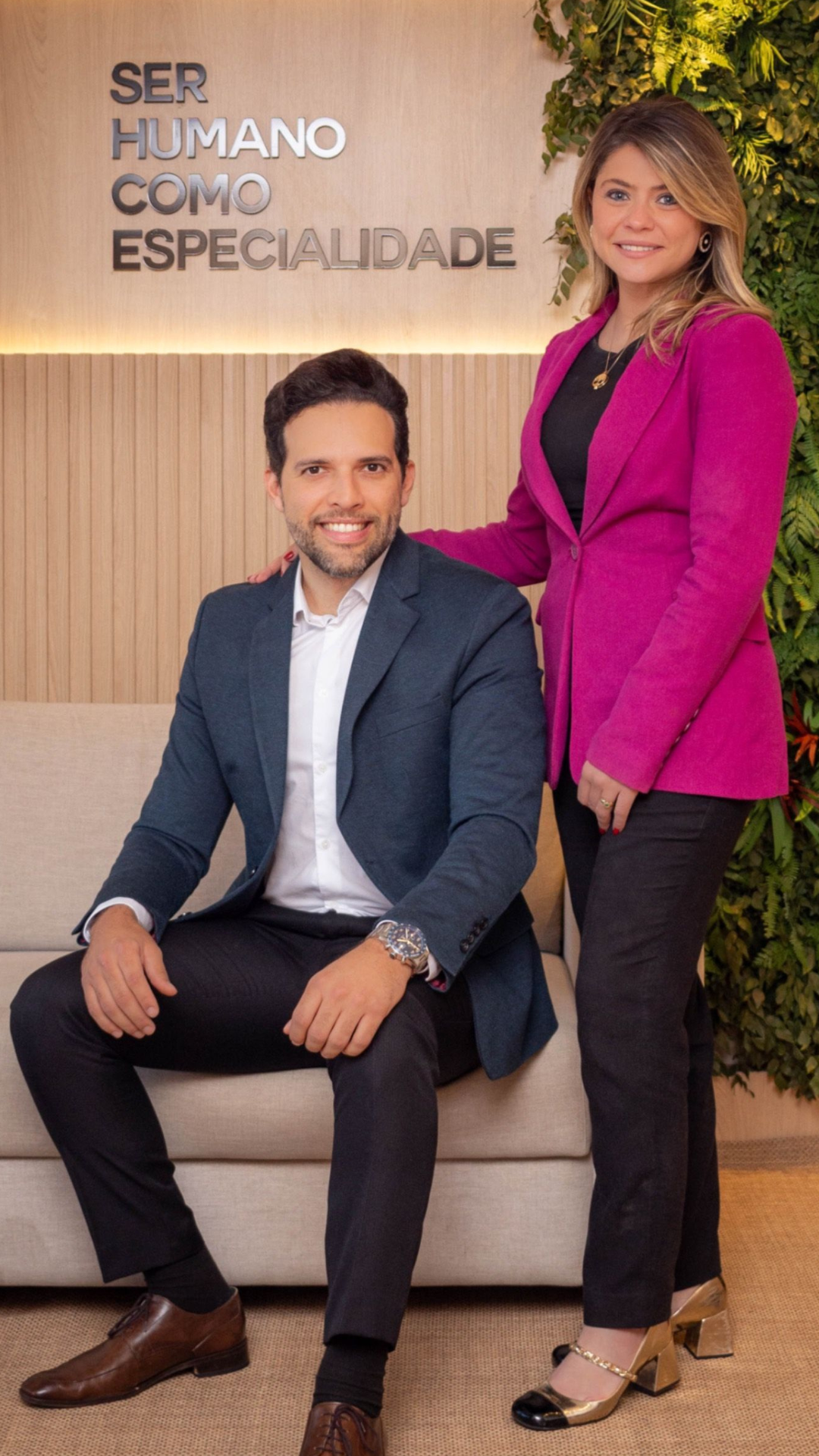Dois profissionais, medicos psiaqutras, elegantes, bonitos, confiantes, mostram autoridade, em um ambiente de escritório, uma sentada e a outra em pé, com um fundo de vegetação e uma parede de madeira com a frase "SER HUMANO COMO ESPECIALIDADE".