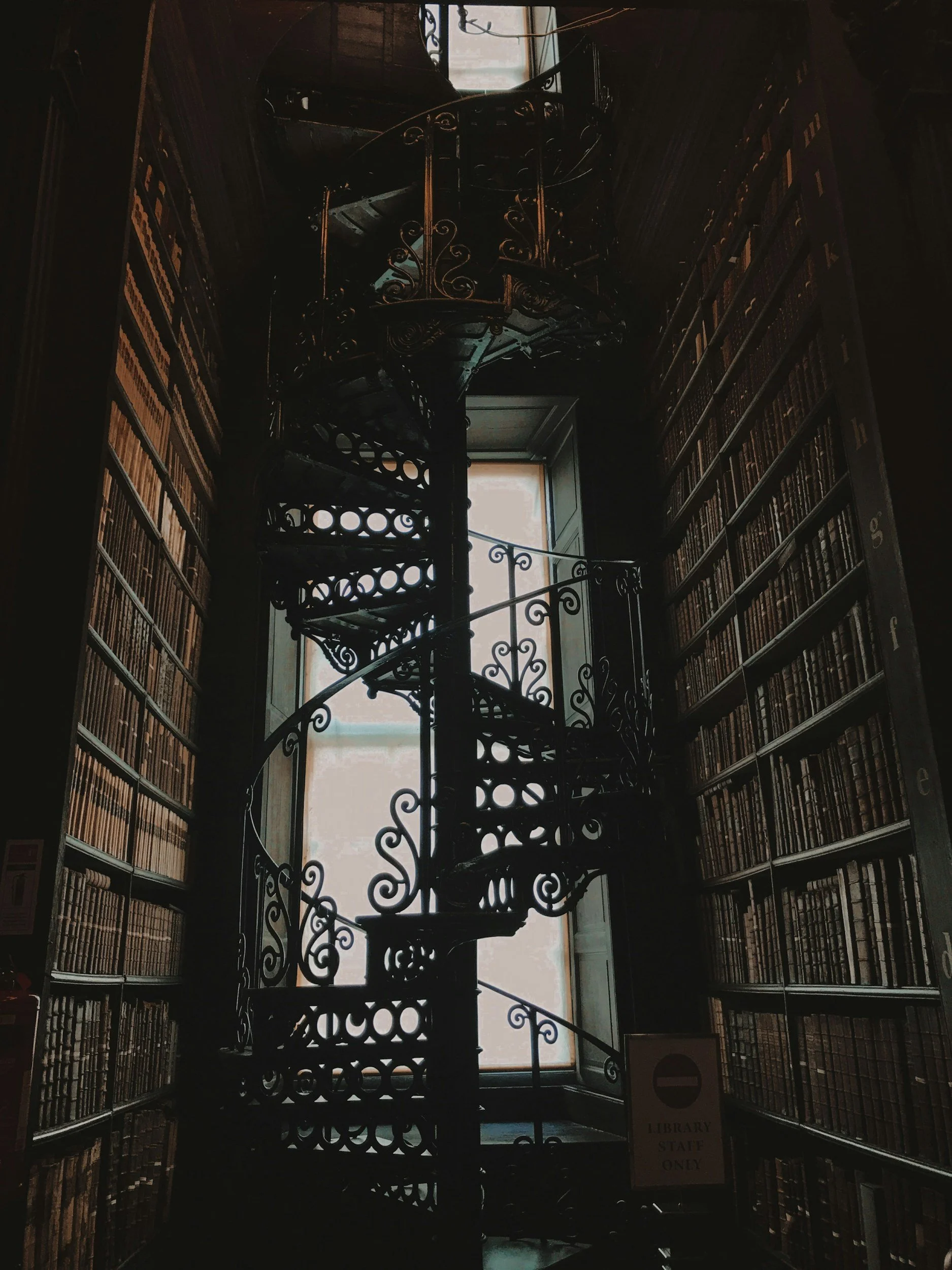 Interior of a library with tall wooden bookshelves on both sides filled with books. There is a spiral staircase with ornate black iron railing in the middle, leading up to a second level. A large window at the back allows natural light to fill the space.
