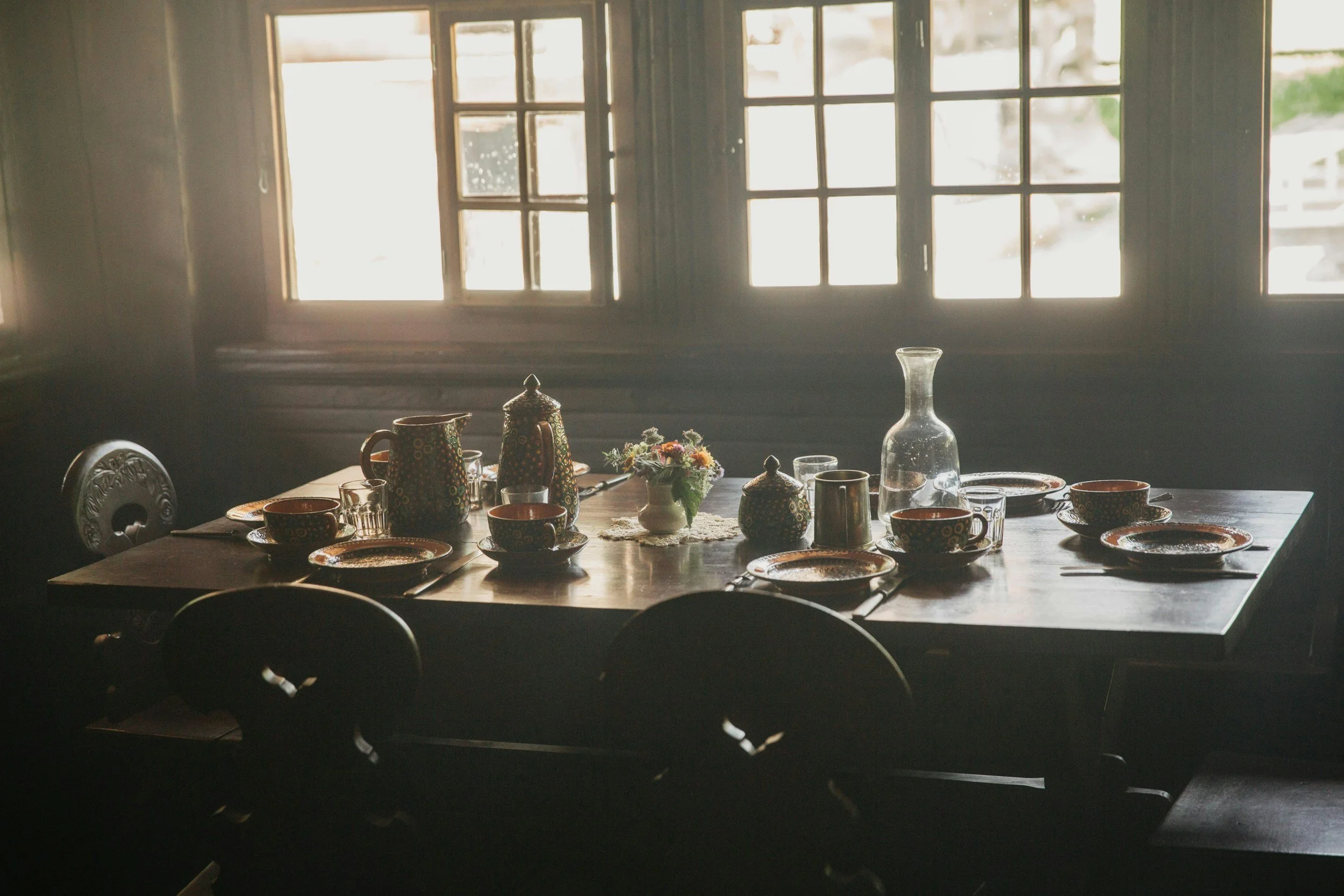 A dimly lit dining room with a wooden table set with various cups, bowls, a pitcher, and a small flower arrangement. Bright light streams through multiple-pane windows in the background.