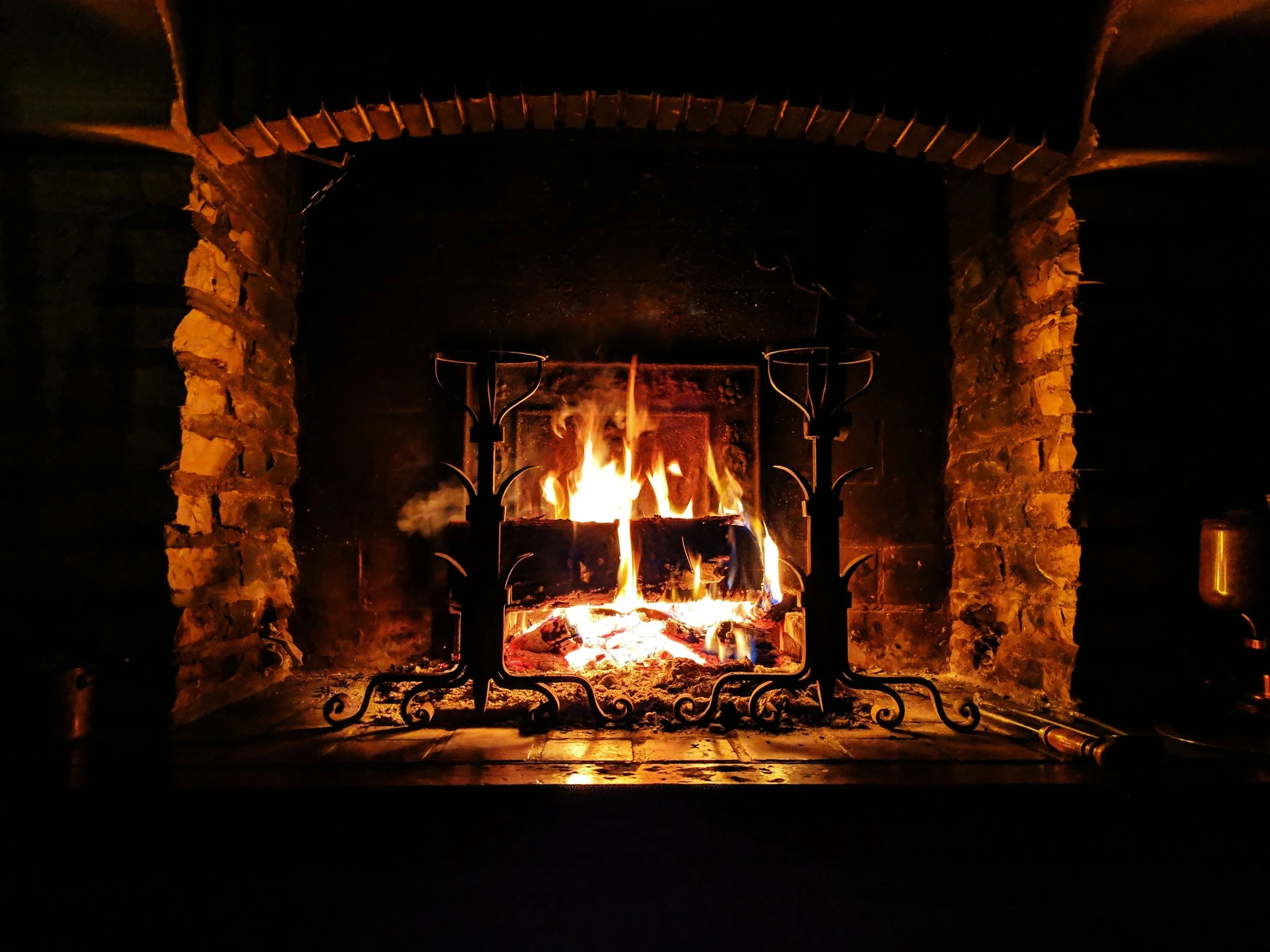 A lit fireplace with a stone surround and a metal grate, flames flickering and burning logs inside.