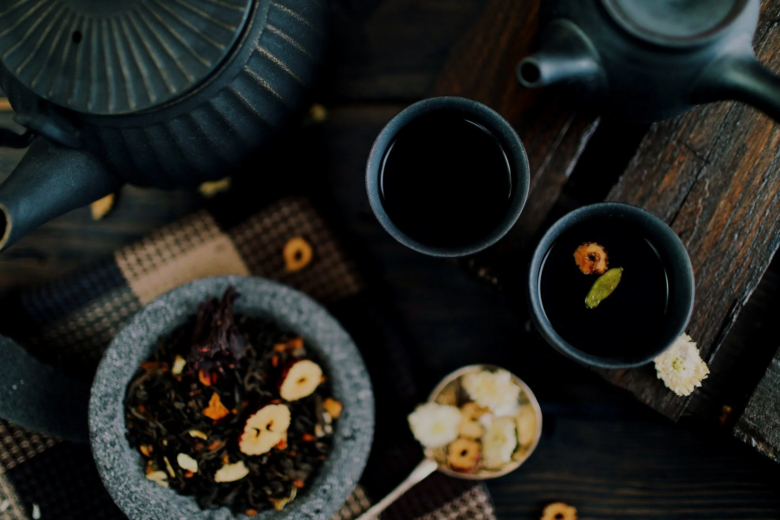 Two tea kettles and multiple cups of tea, along with bowls of cookies and snack items on a dark wooden table.