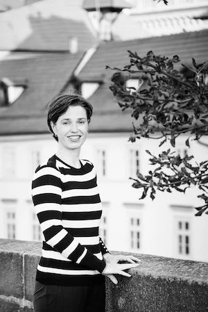 A woman with short hair wearing a striped long-sleeve shirt stands outdoors on a balcony with a cityscape in the background.