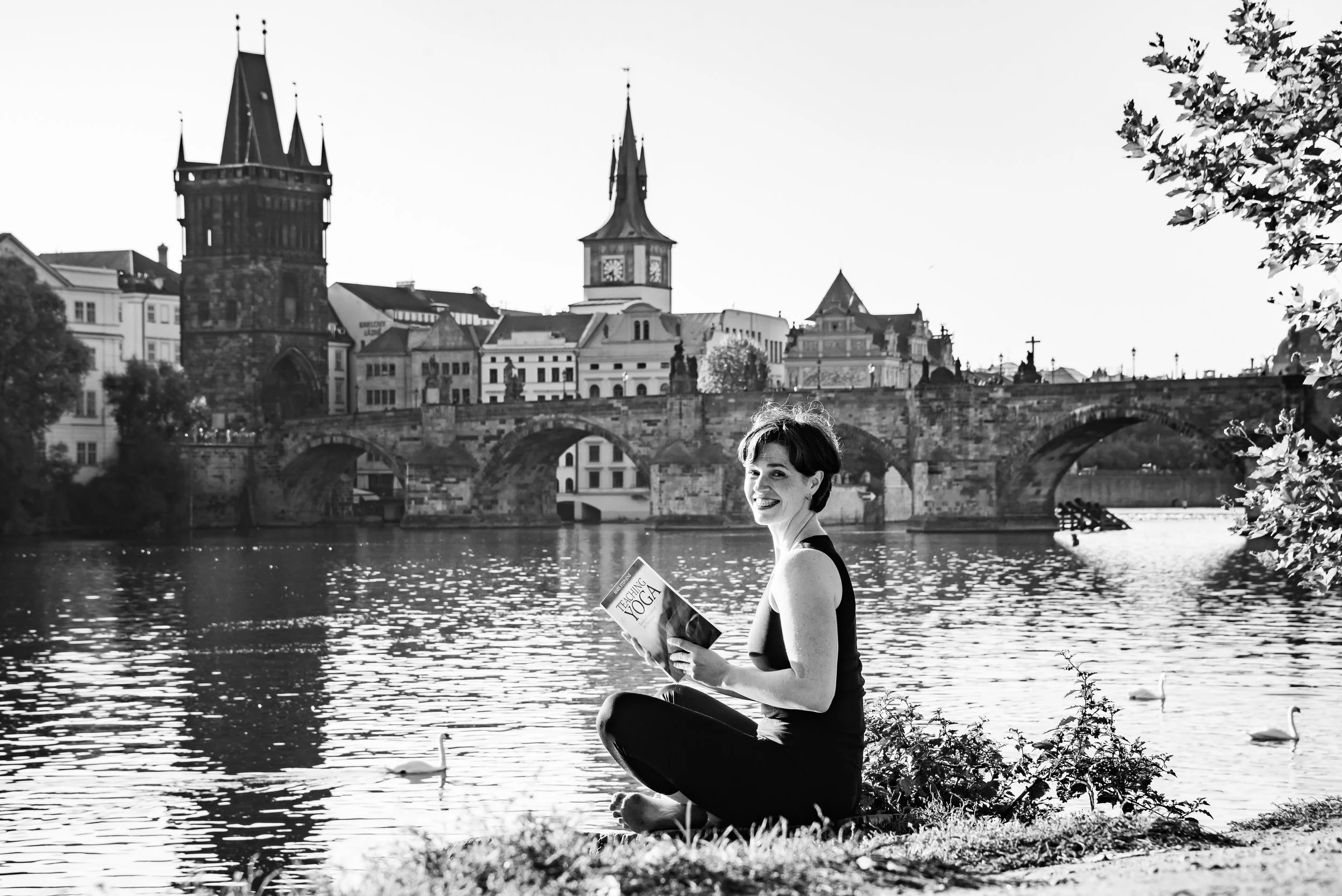 A woman sitting cross-legged by a river, reading a yoga magazine, with historic buildings and a bridge in the background, black and white photo.