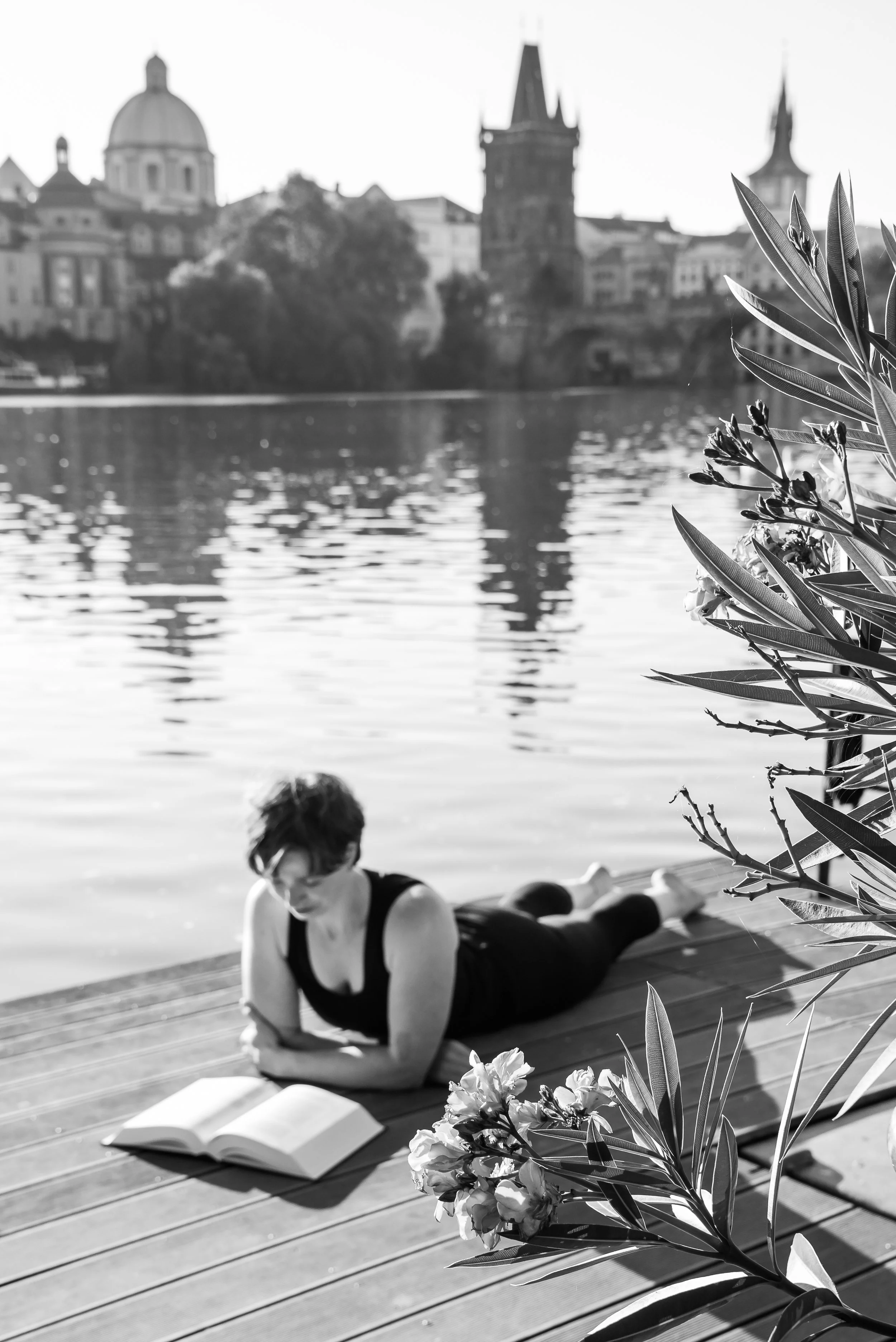 A woman with short hair and glasses is lying on her stomach on a wooden dock, reading a book, near a body of water with a city skyline including a domed building and a tower in the background. Flowers are in the foreground.
