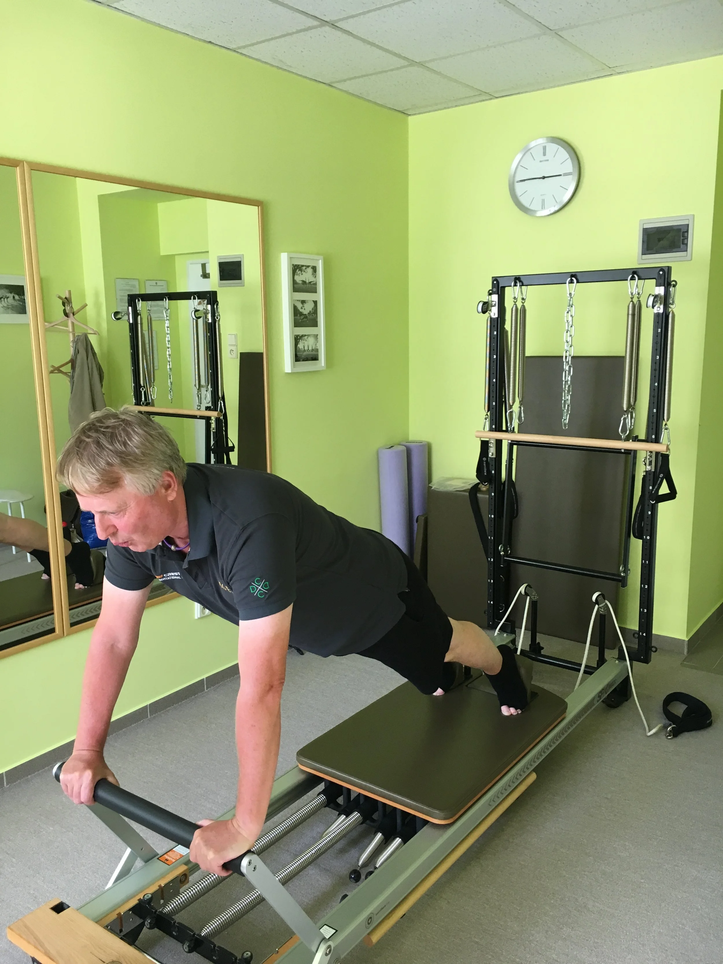 A man in black athletic clothing using a reformer Pilates machine in a fitness room painted in light green, with wall mirror, clock, and framed pictures.