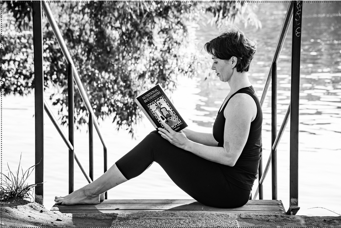 Woman in black athletic wear sitting on a boat dock by the water, reading a book.