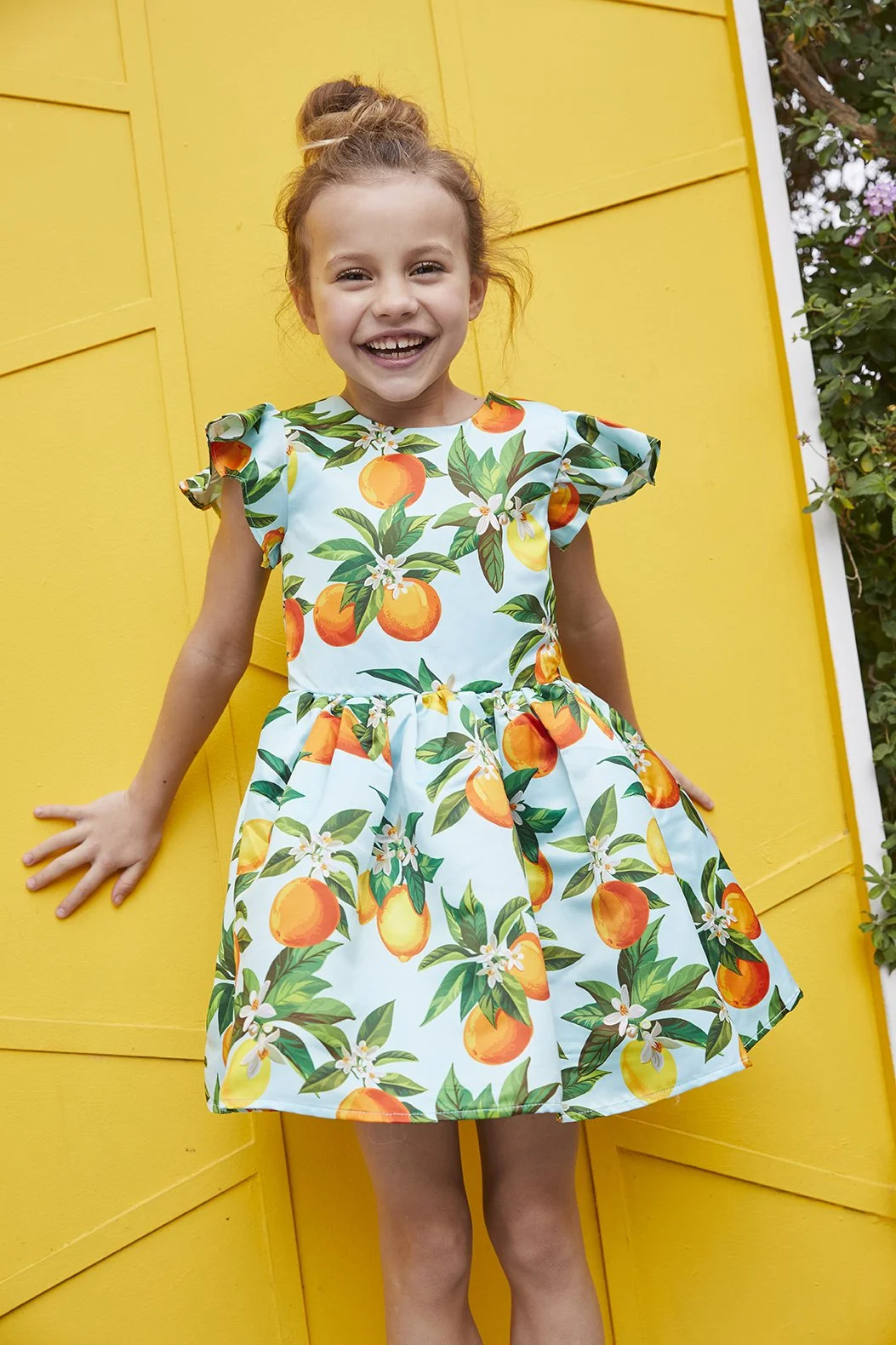 A young girl in a dress with orange and yellow fruit pattern smiling, standing against a bright yellow background, outdoors with greenery on the side.