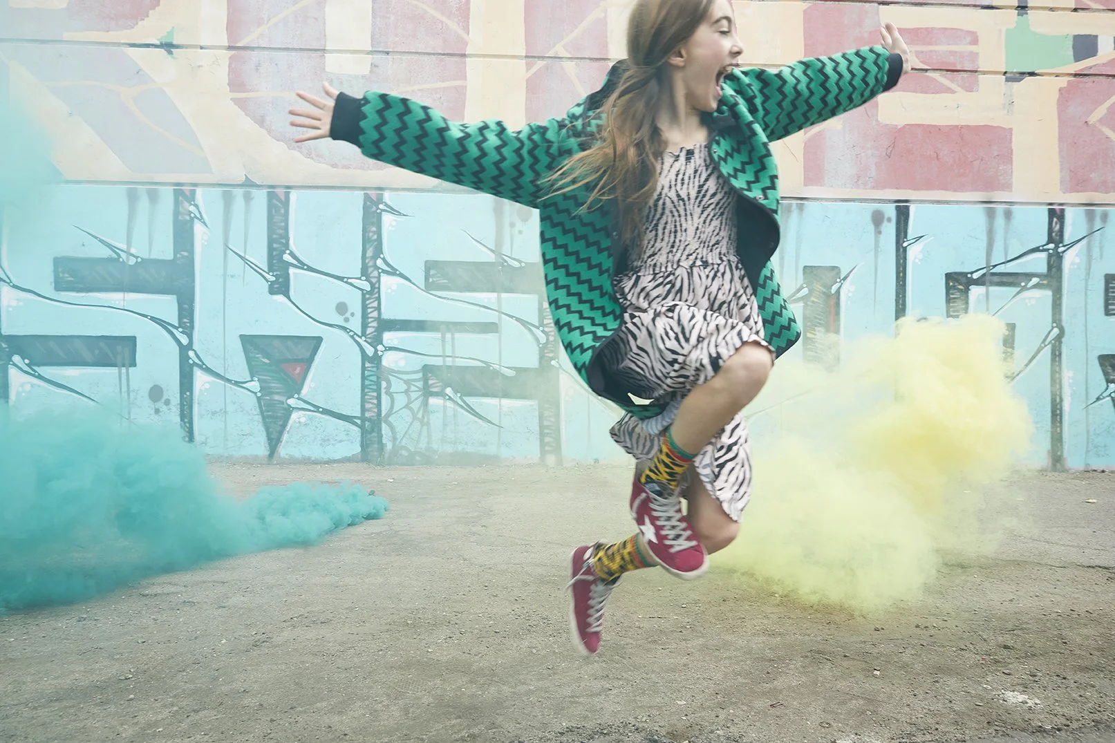 A young girl in a zebra print dress, red sneakers, and colorful socks is jumping in the air with her arms outstretched and her mouth open in excitement. She is outdoors in front of a graffiti-covered wall with blue and pink tones, surrounded by blue 
