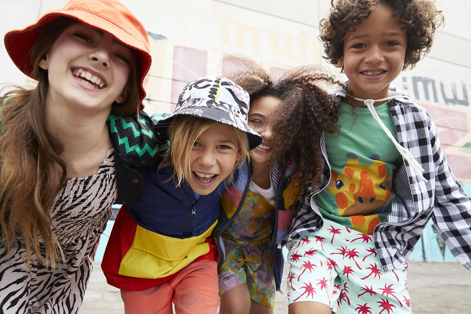 A group of five children smiling and leaning into each other at a colorful outdoor mural, dressed in casual, vibrant clothing.