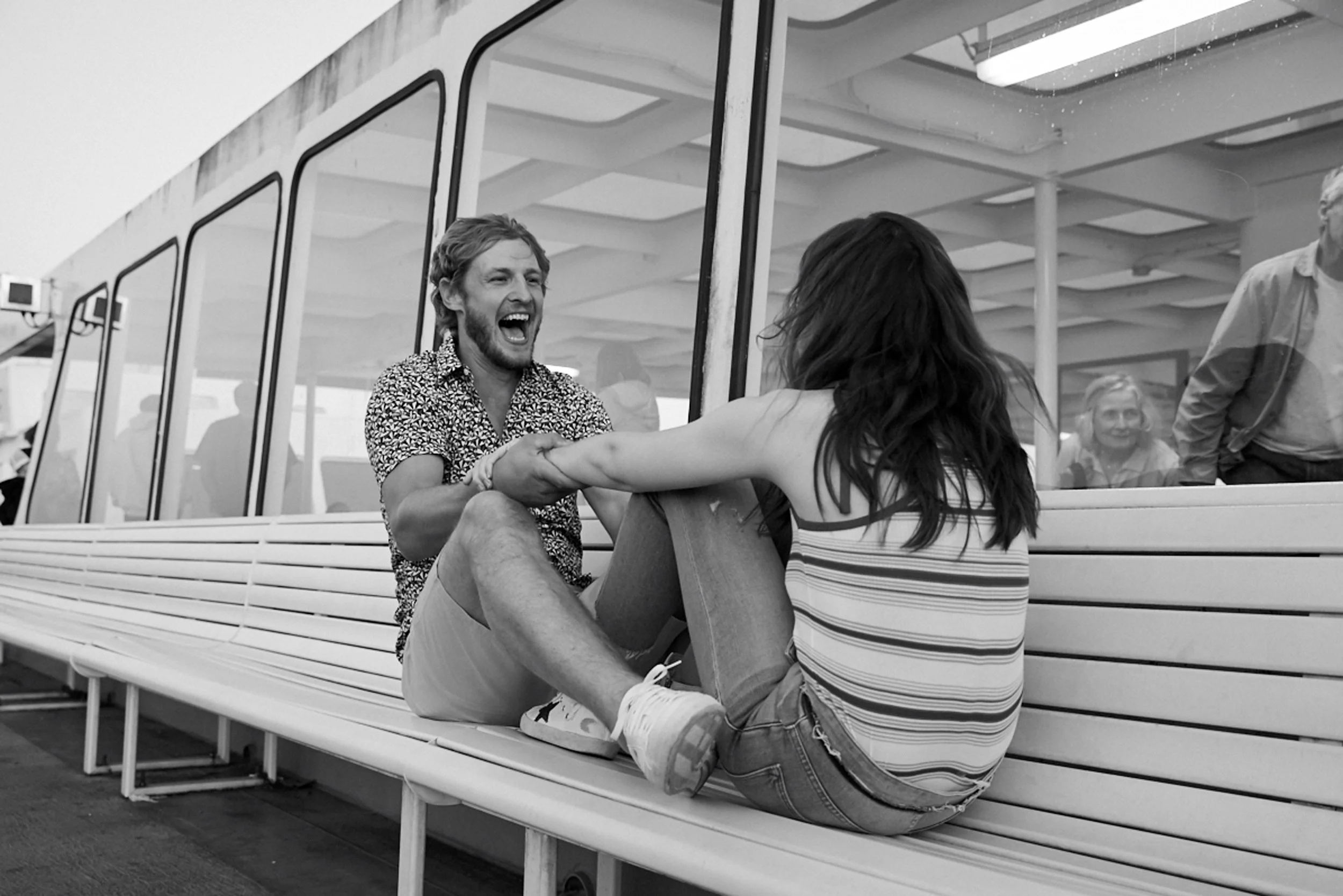 A man and woman sitting on a bench holding hands and smiling, with other people visible through a glass window behind them.