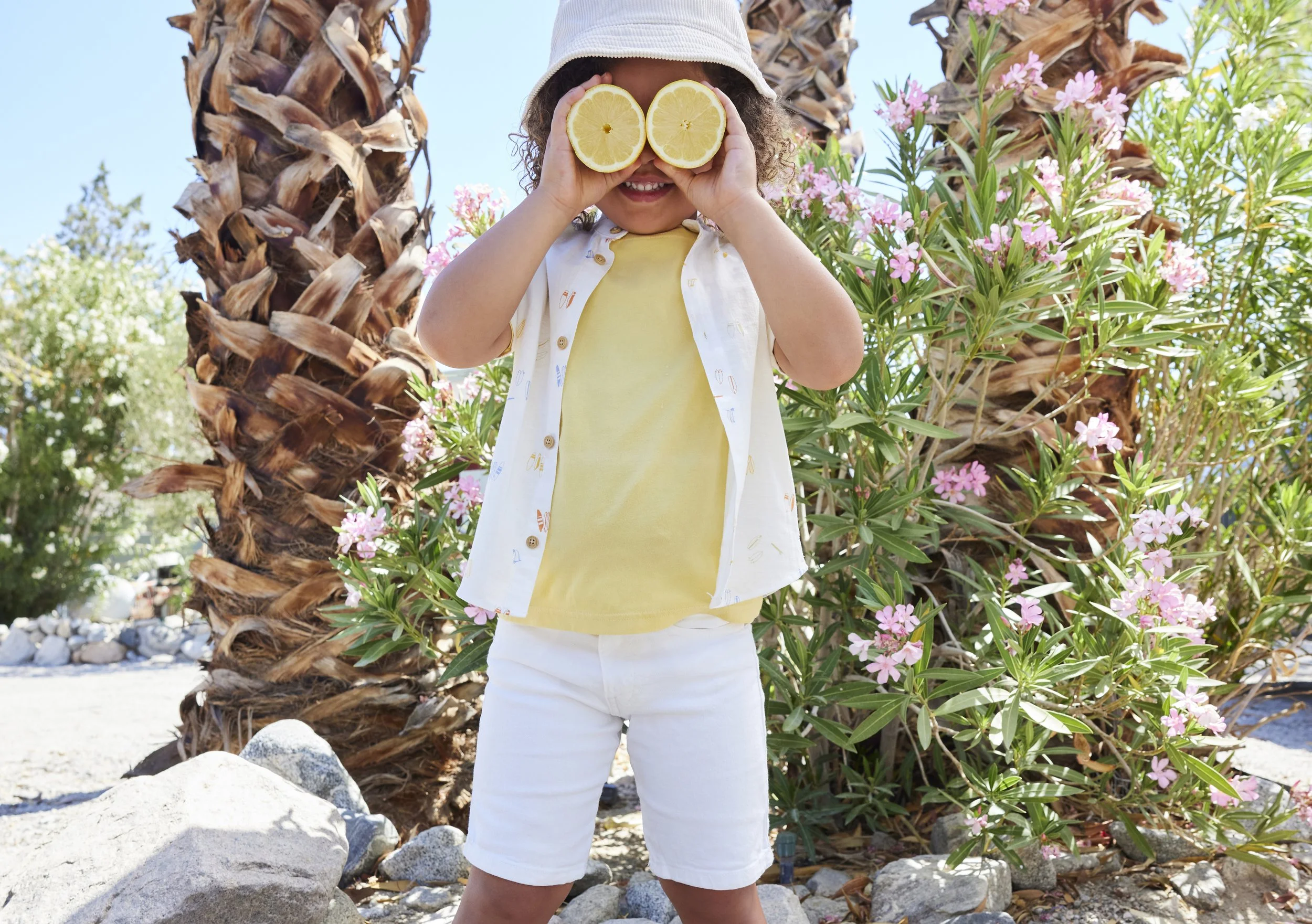 Child outdoors holding lemon slices up to eyes, smiling, among palm trees and pink flowers, wearing a hat, yellow shirt, white shorts, sunny day.