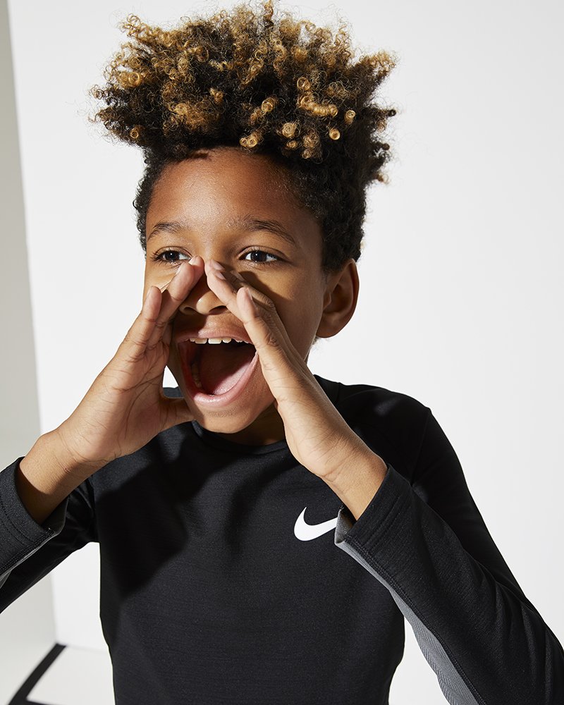 A young boy with curly, light brown and blonde hair, smiling and pretending to shout, wearing a black Nike sports shirt.