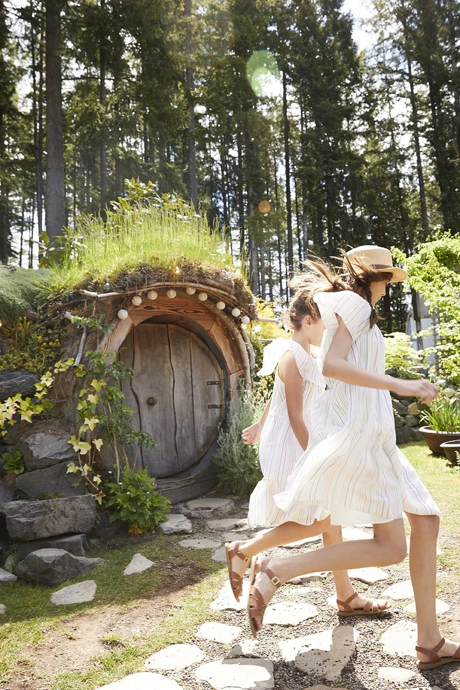 Two girls in white dresses running outdoors near a round wooden door built into a grassy hillside, surrounded by trees.