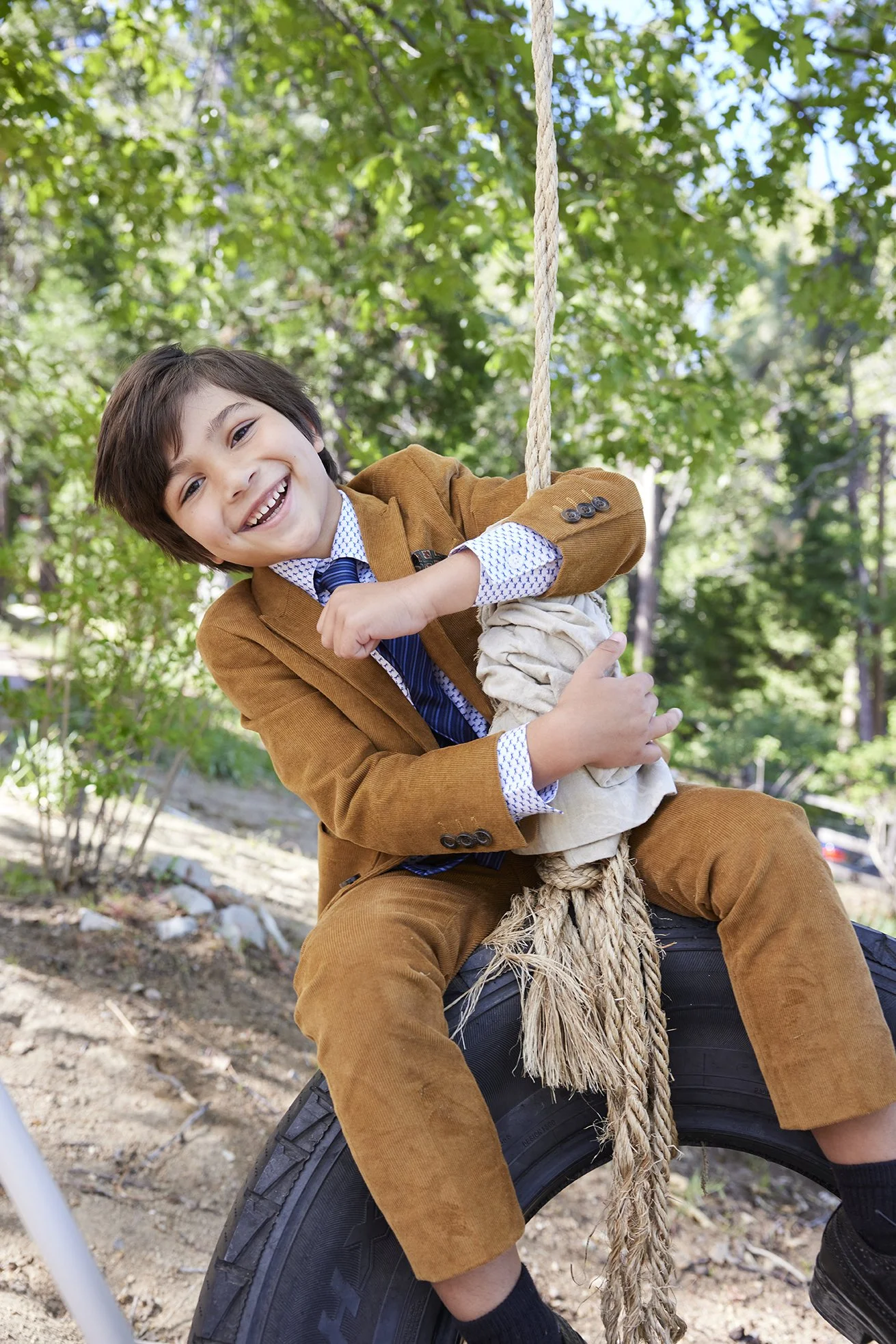 A young boy in a brown suit is smiling and playing on a tire swing outdoors, surrounded by green trees.
