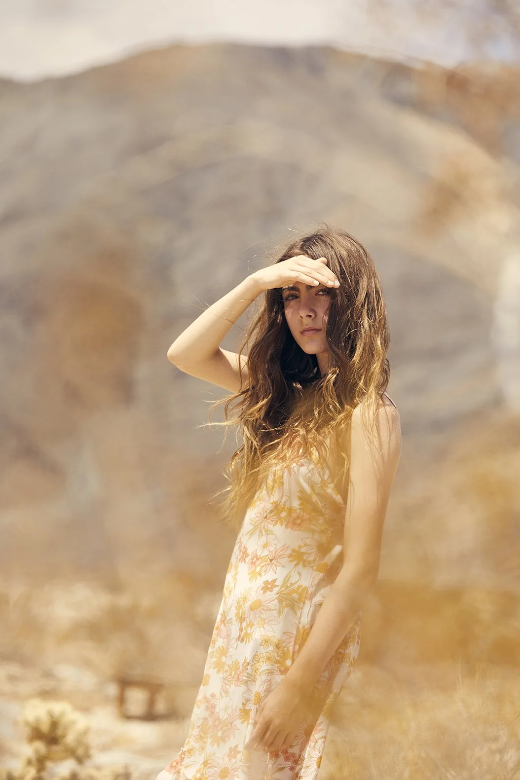 A young woman with long, wavy brown hair wearing a floral dress stands outdoors. She is shielding her eyes from the sun with her hand, and the background features a large, blurred mountain or hill.